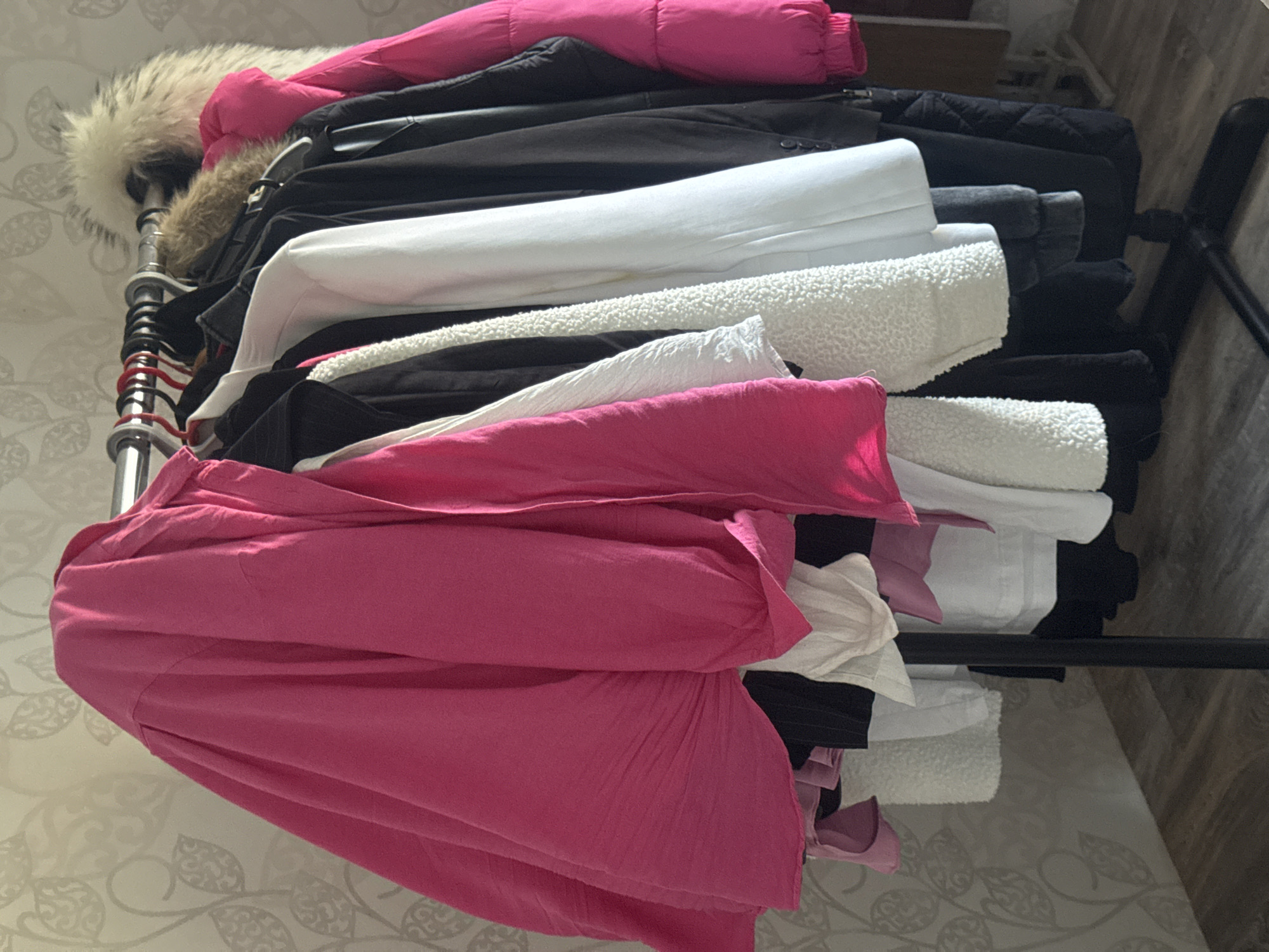 The image shows a clothing rack with several jackets and coats hanging on it. The jackets are of various colors, including pink, black, and white. Some of the jackets have fur trim. The clothing rack is set up in what appears to be a room with a patterned wallpaper and a wooden floor. The lighting in the room is soft, suggesting it may be daytime. The image is a close-up shot, focusing on the clothing and the rack. There are no people visible in the image. The image has been cropped to highlight specific areas of the clothing rack and the garments. The city of Kremenchuk, Ukraine is not depicted in this image.