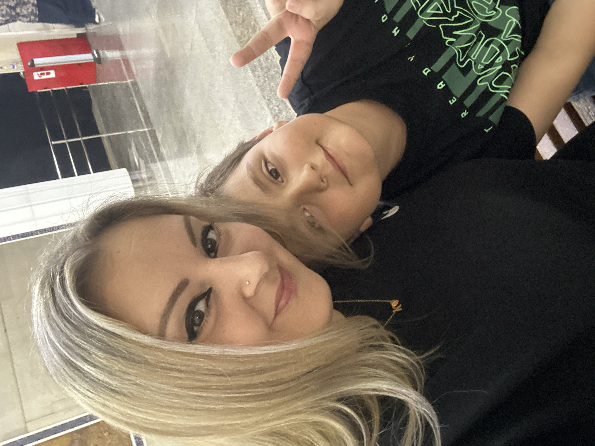 A selfie is taken from a low angle, featuring a woman with blonde hair and a young boy. The woman, positioned on the left, is wearing a black top and a gold necklace with two small letter pendants. She has a nose piercing and is wearing winged eyeliner. The boy, on the right, is wearing a black t-shirt with green lettering and is making a peace sign with his right hand. His left arm is resting on the woman's shoulder. They are indoors, with a blurred background that includes a polished floor, metal railings, and a red fire alarm box. The overall lighting suggests an indoor, possibly public, space. The specific location in Tashkent, Uzbekistan cannot be determined from the visual information provided.