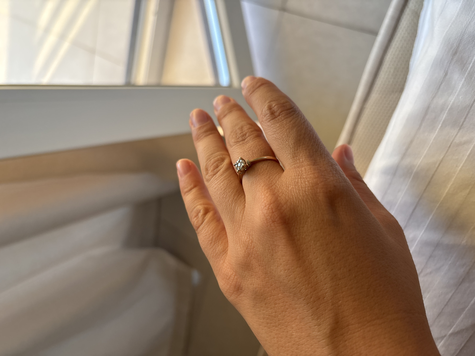 A close-up, eye-level shot shows a person's left hand with a gold ring adorned with a square-shaped diamond, positioned on their ring finger. The hand is relaxed, with fingers slightly spread. The background consists of white curtains on the left and a blurred view of a window with natural light filtering through on the right. The setting appears to be indoors. The image does not provide any information related to Ldzaa, Abkhazia.