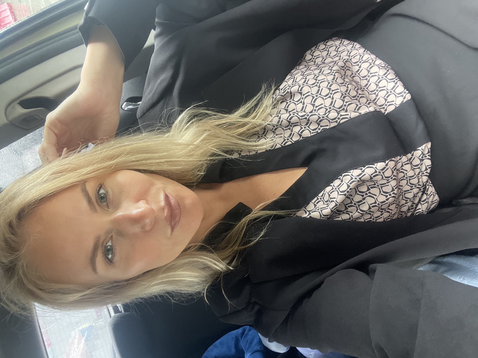 A selfie of a blonde woman with green eyes is taken inside a car. She is wearing a black blazer over a top with a pattern of interlocking shapes. The car interior is visible around her. The specific location in Sochi, Russia, is not discernible from the image.