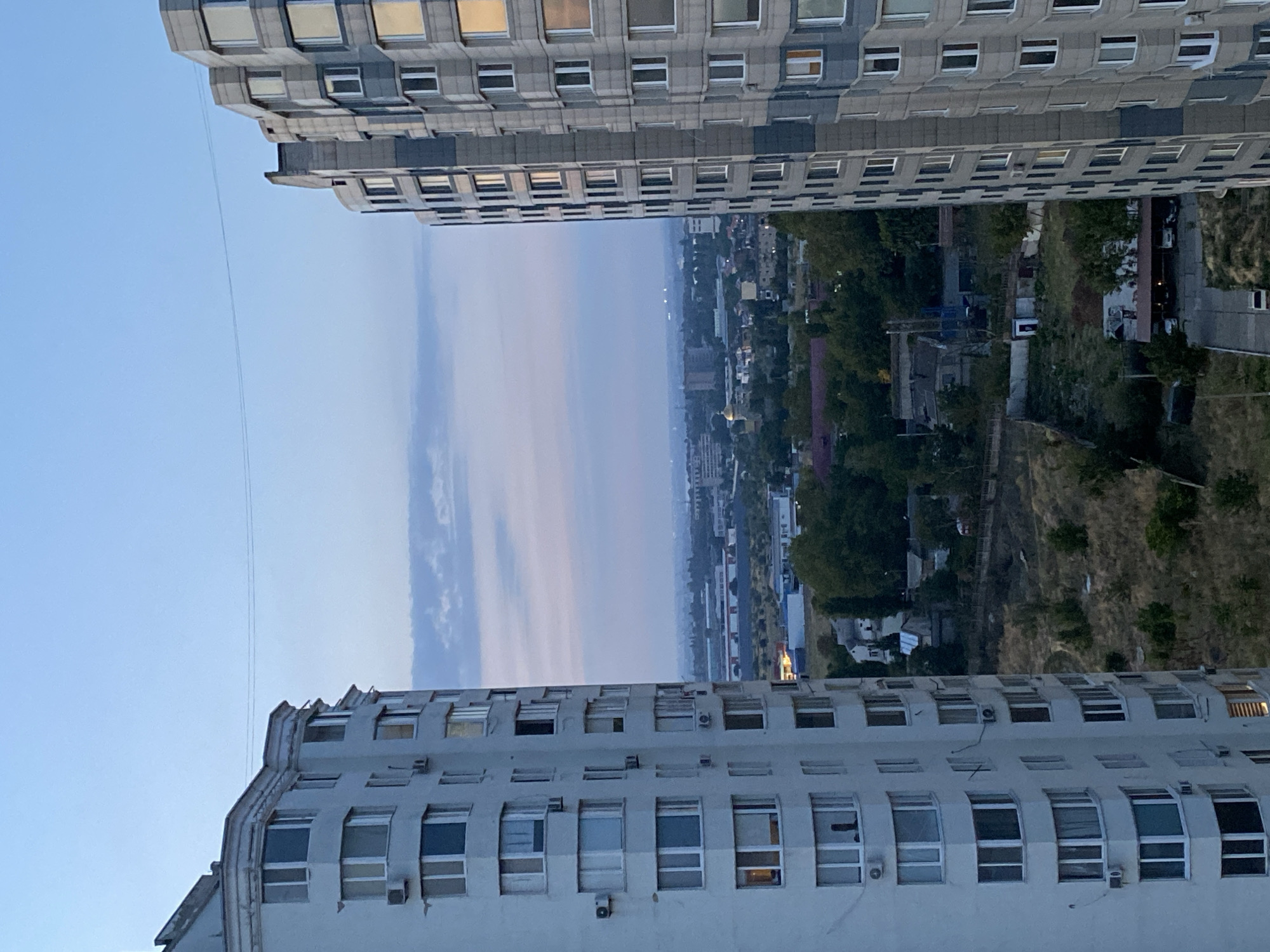 This is an eye-level, medium shot of two tall apartment buildings in Chornomorsk, Ukraine, framing a view of the city and sky at dusk. The building on the left is white and has many windows. The building on the right is gray and has a more geometric facade with multiple angled window panes. In the middle ground, there are smaller buildings, trees, and a golden-domed church. The background shows a hazy cityscape. The sky is a pale blue with wispy clouds.