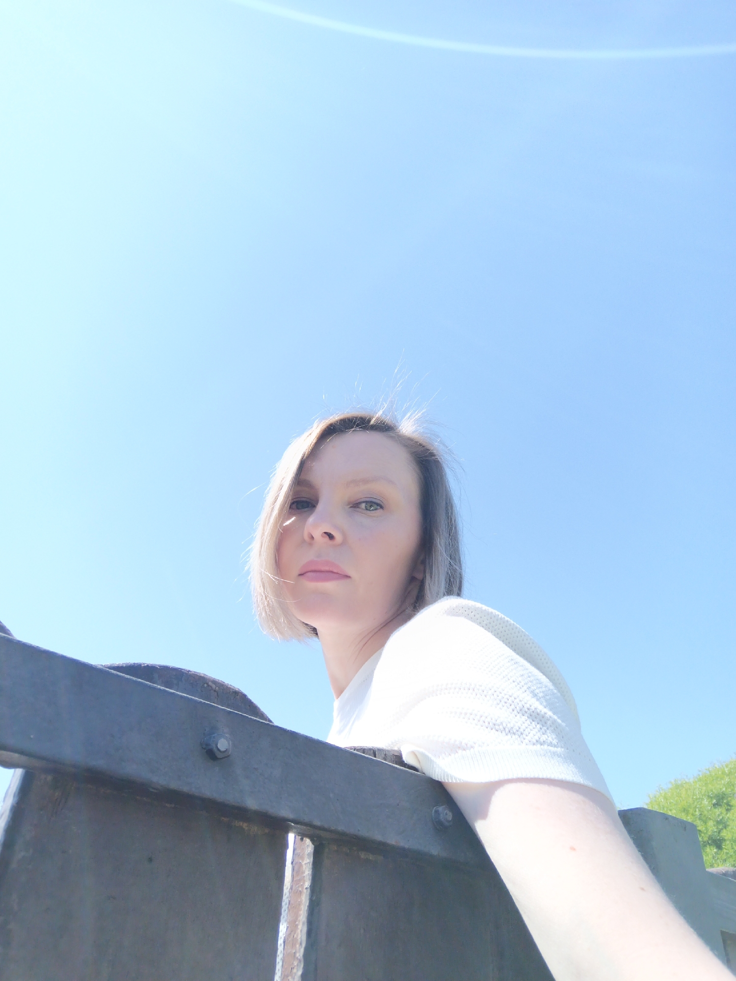 A woman with short, blonde hair is positioned in the lower half of the image, looking directly at the camera. She is wearing a white shirt. The background is a clear, blue sky with a visible light flare. A metal railing or barrier is in the foreground. To the right of the railing is some green vegetation. No specific location within Chaikovsky, Russia is definitively evident.
