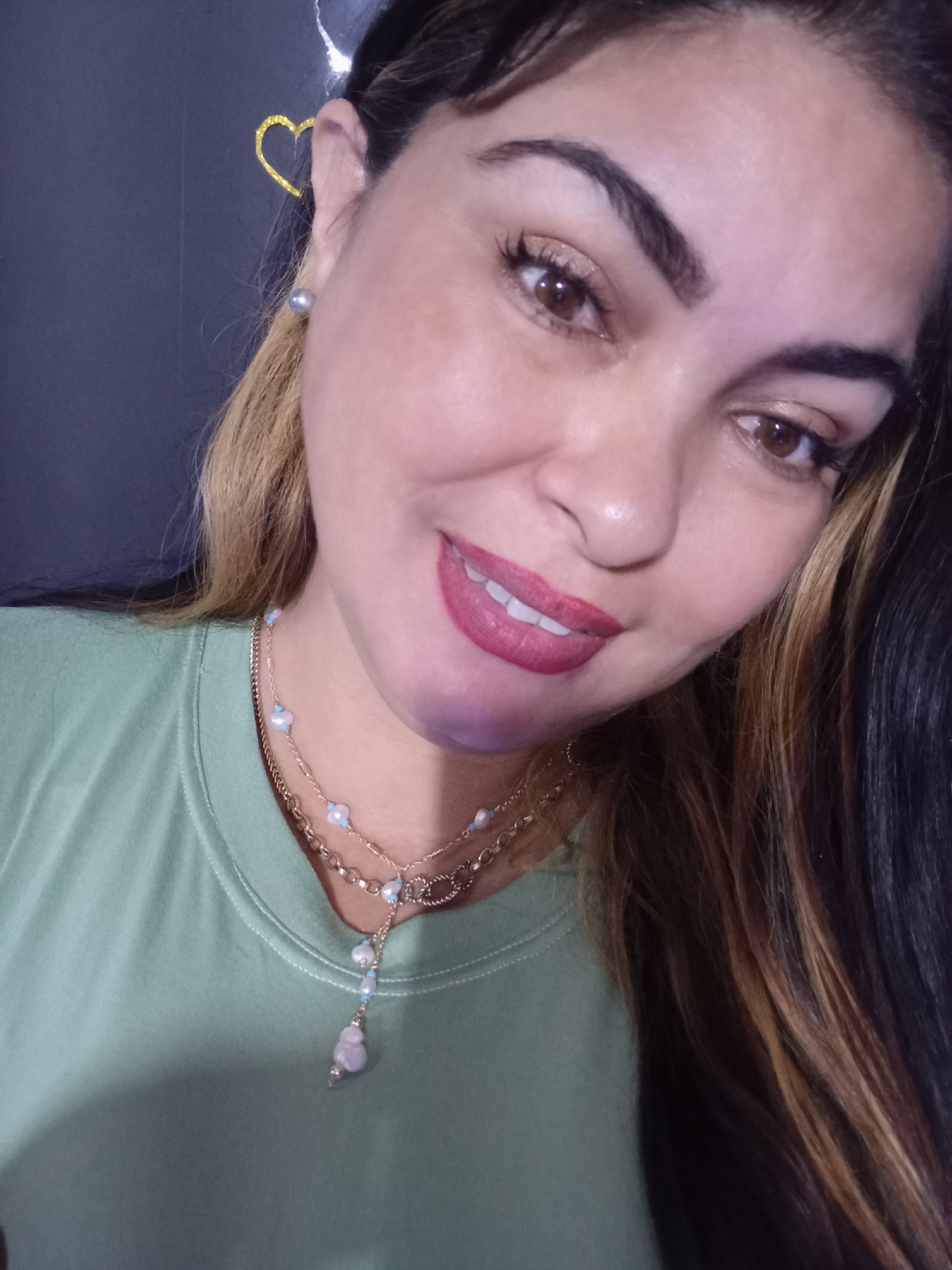 This close-up portrait features a woman with fair skin, dark brown eyes, and dark, well-defined eyebrows, looking directly at the camera with a wide smile that reveals white teeth. Her lips are painted a deep rose color. Her hair is long, dark brown with prominent light brown/blonde highlights, cascading over her right shoulder. She wears small, greyish pearl stud earrings, one visible on her right earlobe, and a layered golden necklace. The lower layer of the necklace is adorned with small, light blue beads and a 'Y' drop featuring a cluster of similar beads and a larger, light purple stone pendant. She is dressed in a light green crew-neck top. The background is a solid dark color, possibly black, with a golden glitter heart-shaped decoration visible behind her upper left side. The scene is located in the city of Juan Griego, Venezuela.