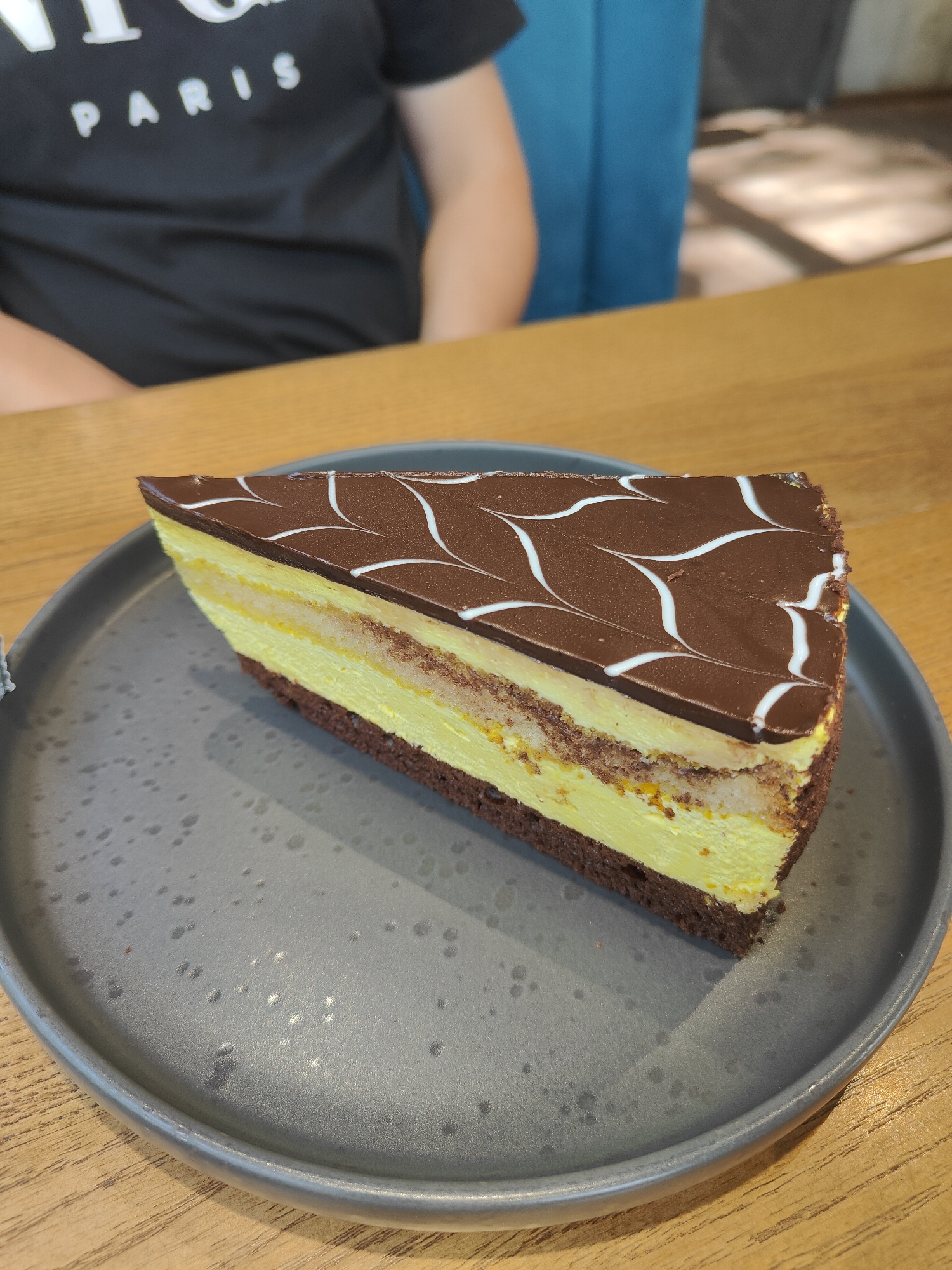 A slice of layered cake sits on a gray, speckled plate on a wooden table. The cake has a dark brown base, followed by a bright yellow layer, a thin crumb layer, another yellow layer, and is topped with dark chocolate icing decorated with white swirled lines. In the background, a person wearing a black t-shirt with white text is visible, seated at the table. The setting appears to be indoors, possibly a cafe or restaurant, with a blue chair and natural light suggesting it's daytime. The overall mood is casual and relaxed, focused on the dessert. The text on the t-shirt is partially visible, reading "PARIS".
