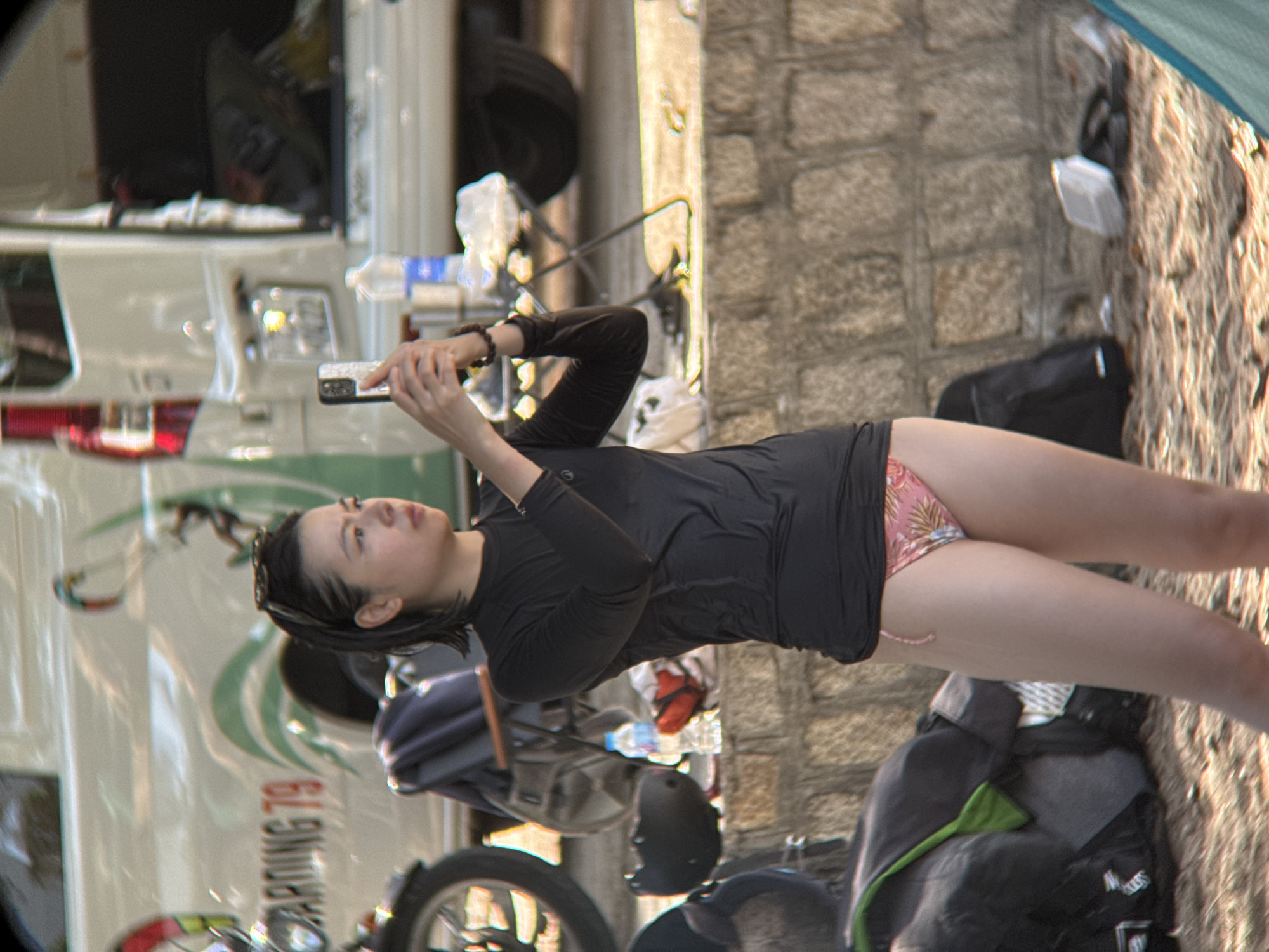 The image captures a woman standing on a sandy area, possibly a beach or park in Nha Trang, Vietnam. She is wearing a black long-sleeved rash guard and a floral-patterned bikini bottom. The woman is holding a smartphone in her right hand, seemingly taking a photo or video. In the background, a white van with text "BOARDING 79" is visible, along with a motorcycle. Other objects like a backpack, a helmet, and a bottle of water are scattered around the scene. The environment appears to be outdoors, with a stone wall in the mid-ground.