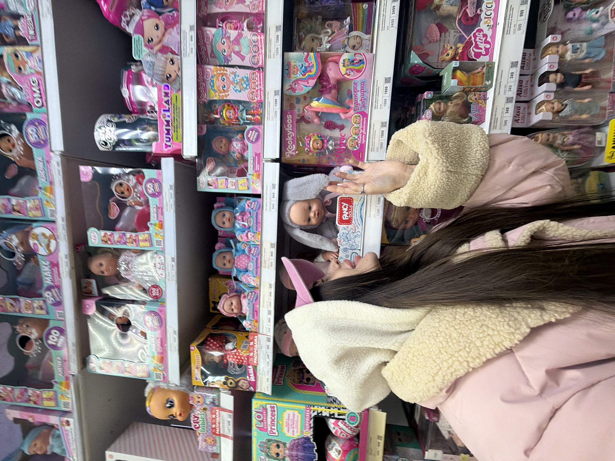 The scene is set within a retail toy aisle in Saint Petersburg, Russia. A female subject, viewed from her right side and rear, occupies the foreground. She possesses long, dark hair and is clad in a light pink puffer jacket with an attached, cream-colored fleece-lined hood. Her gaze is directed towards the right, engaging with the merchandise displayed on shelves. Her right hand is elevated, fingers slightly curled, positioned near a shelf of toys. A ring is present on her left ring finger. The background consists of multiple, well-stocked white retail shelves. These shelves are filled with numerous boxed children's toys, predominantly dolls and doll sets. Visible brands include 'Cry Babies,' 'L.O.L. Surprise! Princess,' 'Kookyloos,' 'Fancy Dolls,' and others, with packaging featuring both Cyrillic and Latin script. White price tags, displaying costs in Russian Rubles, are affixed to the front edge of each shelf, indicating prices ranging from 349 to 2499 Rubles.
