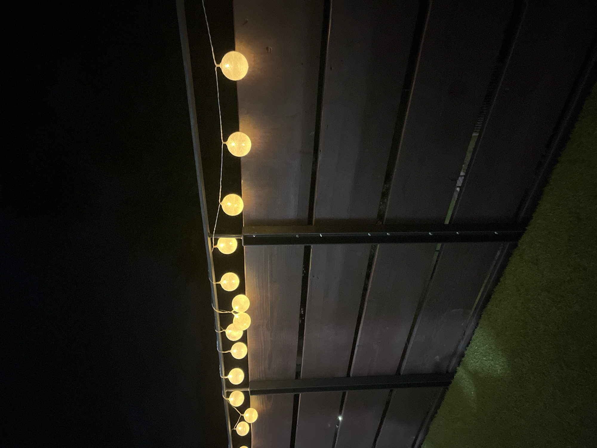 The image depicts a dark outdoor scene with a string of fairy lights illuminating a dark wooden fence at night. The lights are round, glowing yellow, and are suspended by a white cord. The fence is constructed of horizontal wooden planks with vertical supports. Below the fence, there is artificial turf. The sky is completely dark, indicating it is nighttime. There are no people or animals visible in the image. The setting appears to be a backyard or patio area. There is no indication of the city of Garmisch-Partenkirchen, Germany.