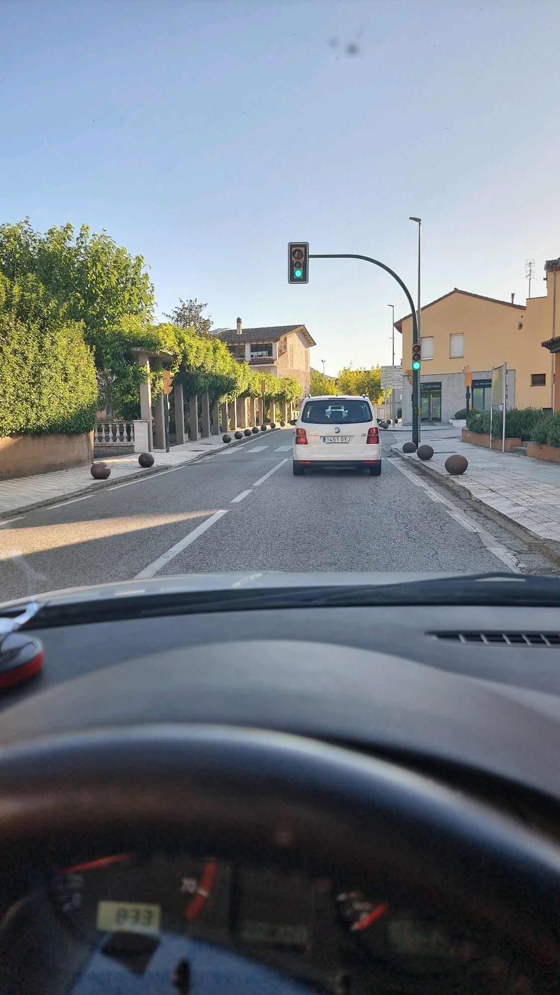 The image depicts a view from inside a car traveling on a street in Sant Joan Les Fonts, Spain. The street is lined with trees and buildings. A white car with license plate "1451 GYJ" is in front of the vehicle from which the photo is taken. Traffic lights ahead are green. The sky is clear and blue. The dashboard of the car shows a digital display with the number "833" and other indicators.