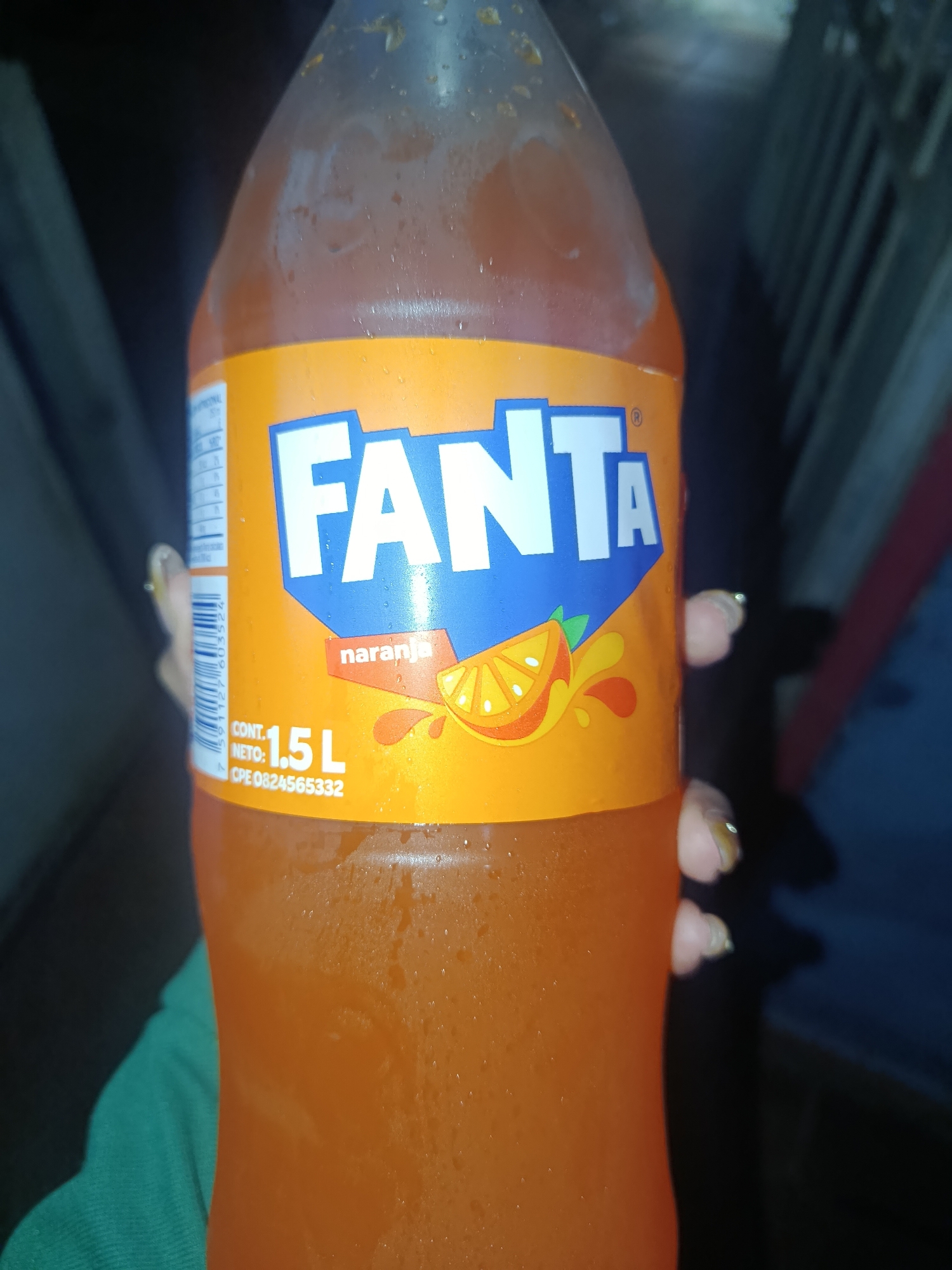 A close-up photograph shows a 1.5 liter bottle of Fanta orange soda being held. The bottle is translucent orange, with an orange label featuring the blue and white Fanta logo and a depiction of a sliced orange. Condensation is visible on the bottle. A hand with painted fingernails is partially visible on the right side of the bottle. A green garment is partially visible on the bottom left corner of the image. The background is dark and indistinct. There is no indication the photo was taken in Juan Griego, Venezuela.
