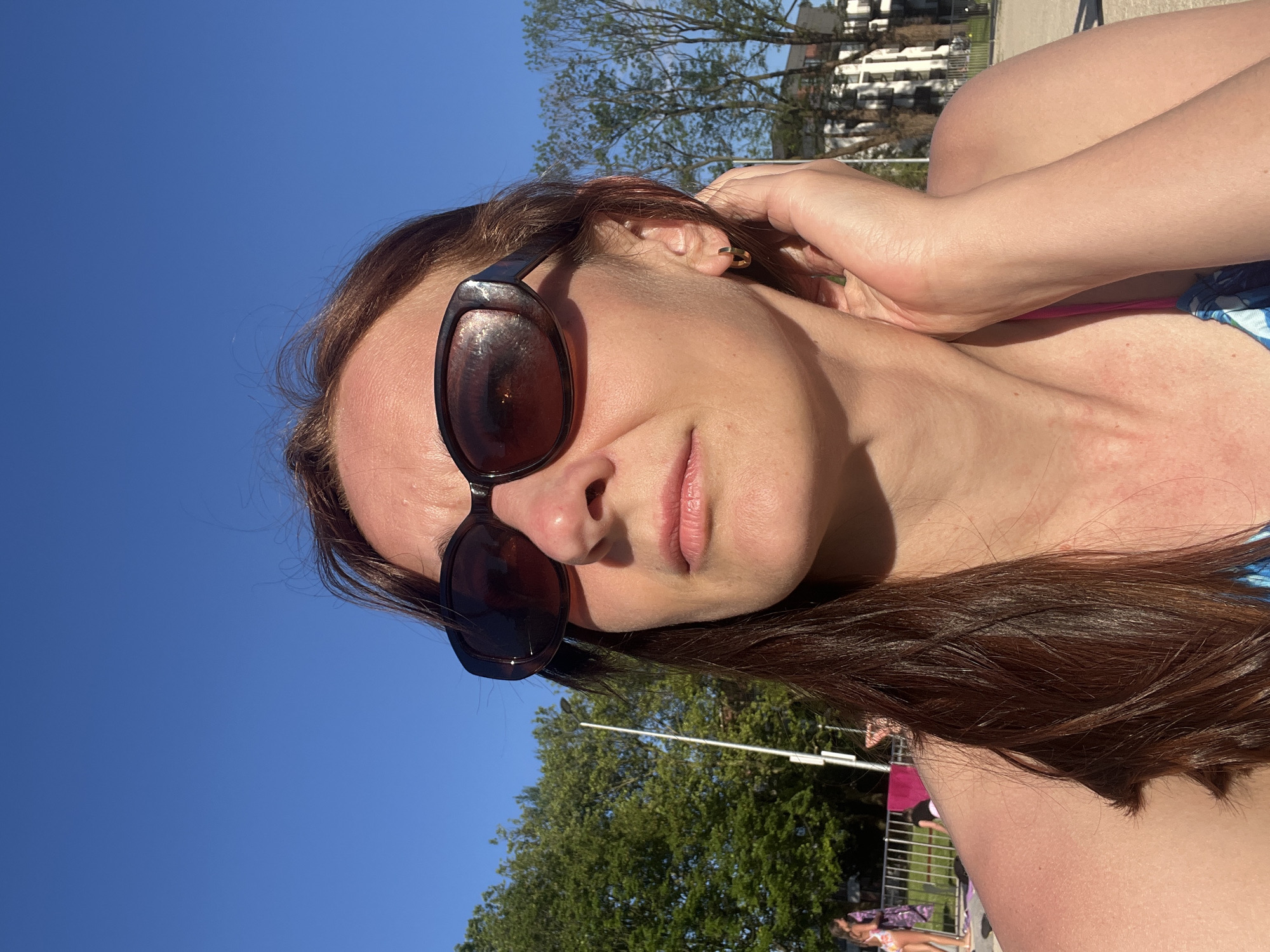 The image shows a woman outdoors on a clear, sunny day. She is looking slightly upward and to her left, wearing dark brown, oval-shaped sunglasses. Her medium brown hair is visible around her face and draped over her left shoulder. A small, gold-colored hoop earring is present in her right ear. Her right hand is gently touching the side of her neck/jawline. She is wearing what appears to be a blue and white patterned top, with a pink strap visible on her left shoulder. Her skin on the shoulders and chest shows some redness. The background features a vibrant blue sky with no clouds. To the left, dense green trees and a tall, white, multi-pronged pole are visible. Further left, behind a silver metal fence, two partially visible individuals are present; one appears to be in a patterned swimsuit. To the right, sparser trees are seen, with light-colored, multi-story buildings in the distance. The scene suggests an outdoor recreational area within an urban or suburban environment.