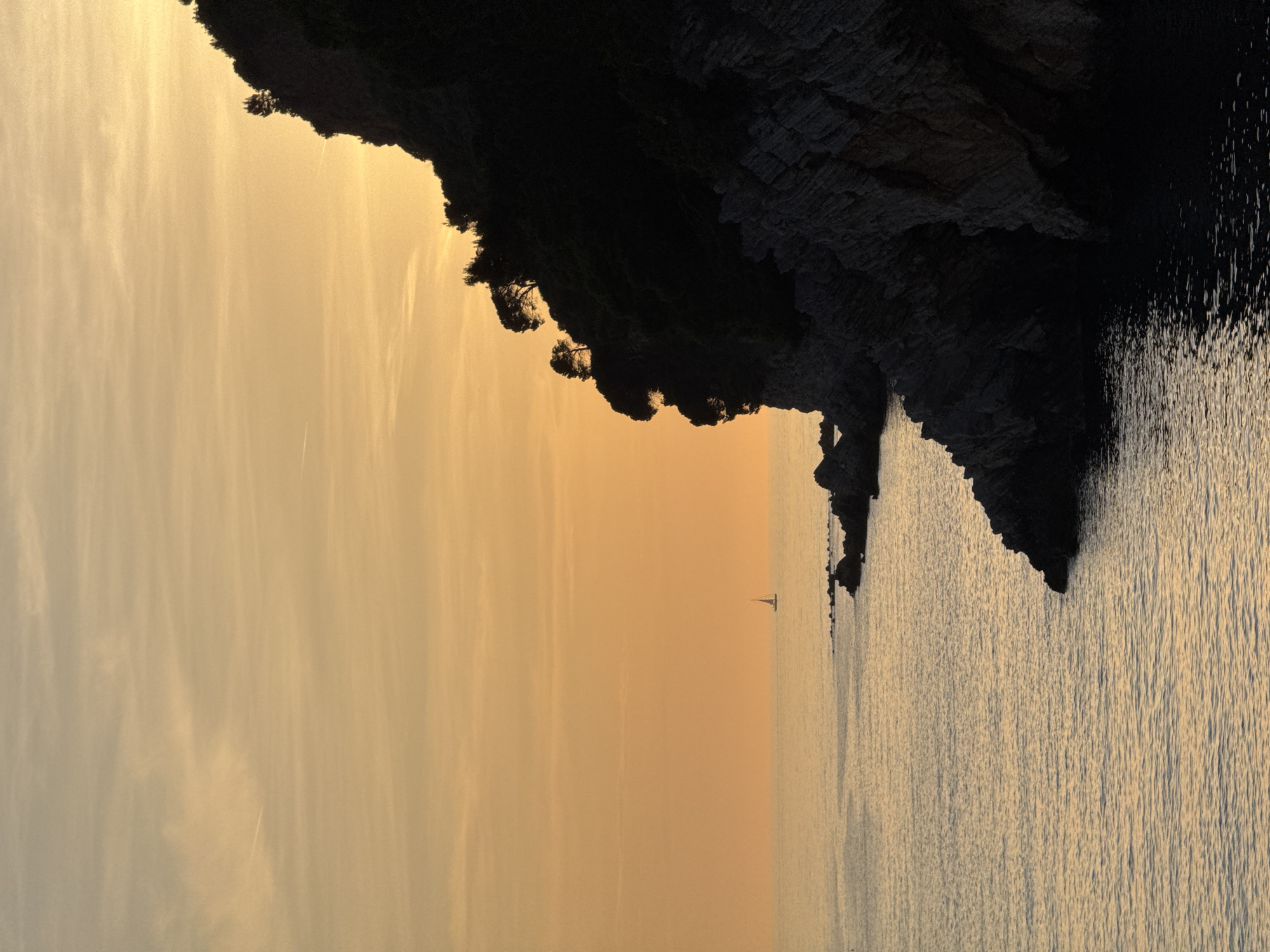 The image depicts a coastal scene in Petrovac, Montenegro. A dark, rocky cliff with sparse vegetation occupies the upper right portion of the frame, descending towards the sea. The sea, reflecting a golden light, fills the lower right and stretches across the bottom of the image. The sky, predominantly yellow and orange in hue, occupies the left side of the frame with faint cloud streaks visible. A single sailboat is visible in the distance near the horizon. The image is oriented sideways.