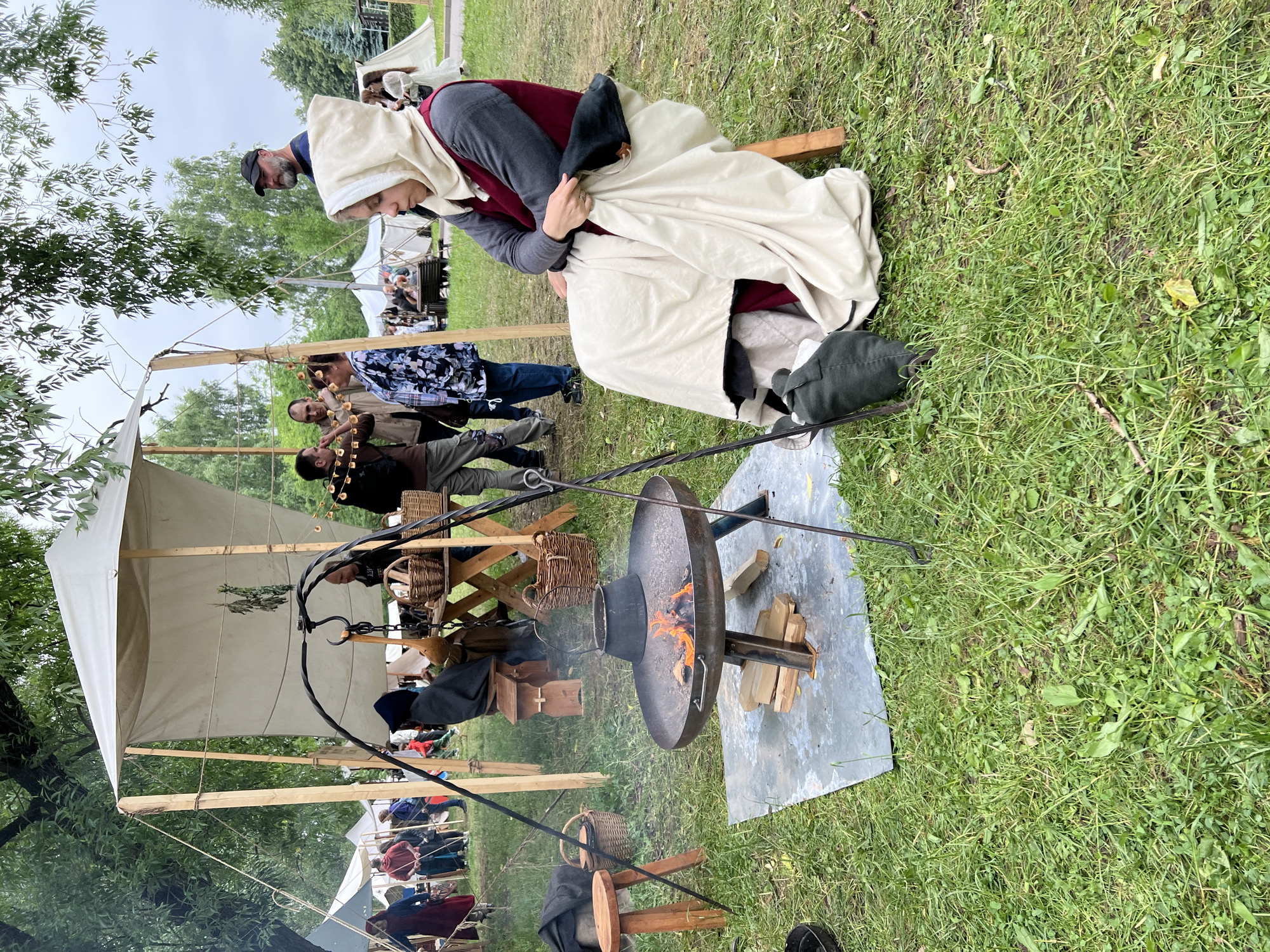The image depicts an outdoor historical reenactment event on a grassy area, possibly in a park. In the foreground, a person dressed in medieval-style clothing, including a hooded garment and a long dress, sits on a wooden stool. The person is looking downwards and appears to be resting. In front of the seated person is a large metal fire pit with a fire burning, and a black pot is suspended above it. Various historical artifacts, such as baskets, wooden stools, and cooking utensils, are scattered around the fire pit and in the background.
Several other people are visible in the mid-ground and background, also dressed in historical attire, suggesting a reenactment of a historical period. Some are standing and conversing, while others are engaged in activities related to the event, such as tending to tents or interacting with visitors. White tents are set up in the background, typical of historical fair or encampment setups. Trees and foliage surround the scene, indicating an outdoor setting.
The specific location in the city of Moscow, Russia, cannot be determined from the visual information provided in the image.