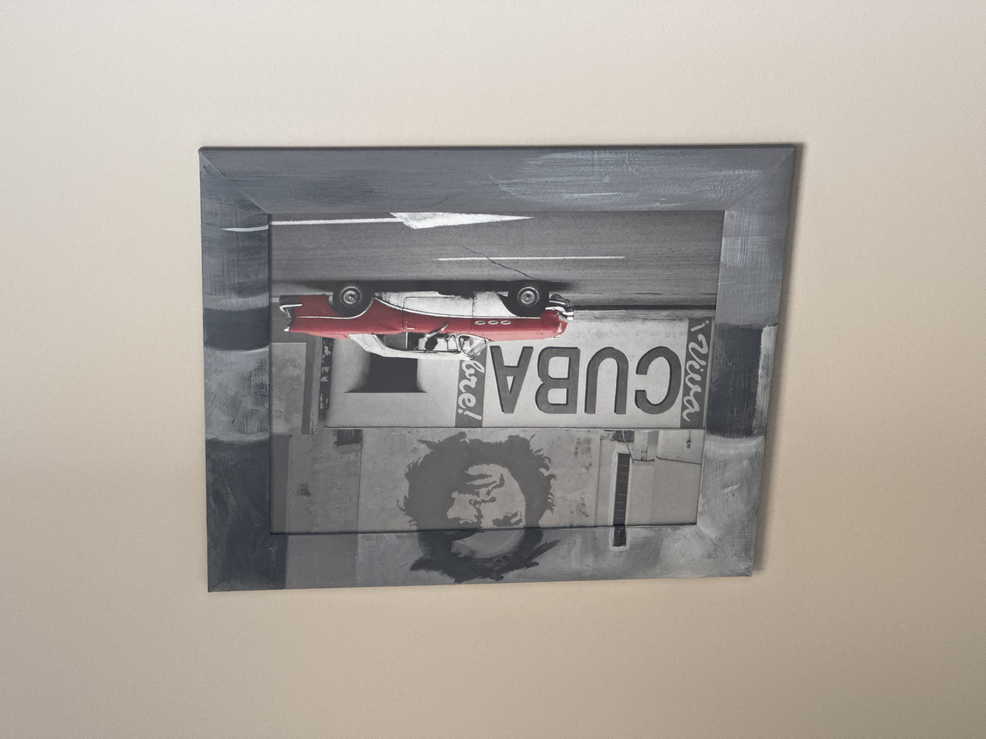The image shows a framed piece of art hanging on a light beige or off-white wall. The art appears to be a mixed-media piece featuring a red and white car, a road with a white arrow, and images related to Cuba, including the word "CUBA" and a portrait of a person. The frame is dark gray with visible brushstrokes. The artwork is mounted horizontally on the wall. There is no indication of the location being Łódź, Poland.