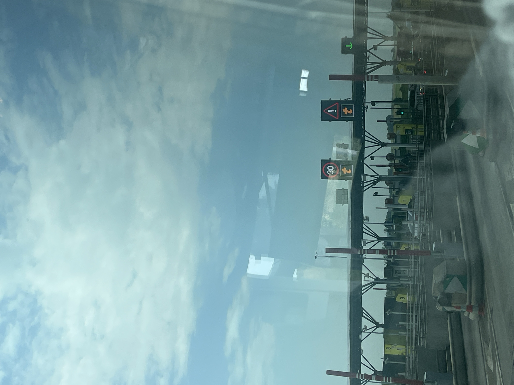 The image depicts a view of a toll plaza, likely from inside a vehicle. The scene is dominated by the overhead structure of the toll booths, supported by dark metal frameworks. Yellow toll booths with numbers such as "8", "9", and "10" are visible. Red and white striped poles mark the lanes. Traffic lights are present, with a green arrow indicating an open lane. Several warning signs are also visible, including one with a red border and an exclamation mark inside a triangle, and another displaying "30" within a red circle, signifying a speed limit. The sky above is a bright, hazy blue with scattered white clouds, suggesting daytime with potentially overcast conditions or haze. The reflection of the sky and clouds is visible on the surface of what appears to be glass, possibly a windshield, creating a slightly distorted and layered perspective of the scene. The overall impression is that of a functional, busy highway environment. There is no visible text other than the numerals on the toll booths and the speed limit sign.