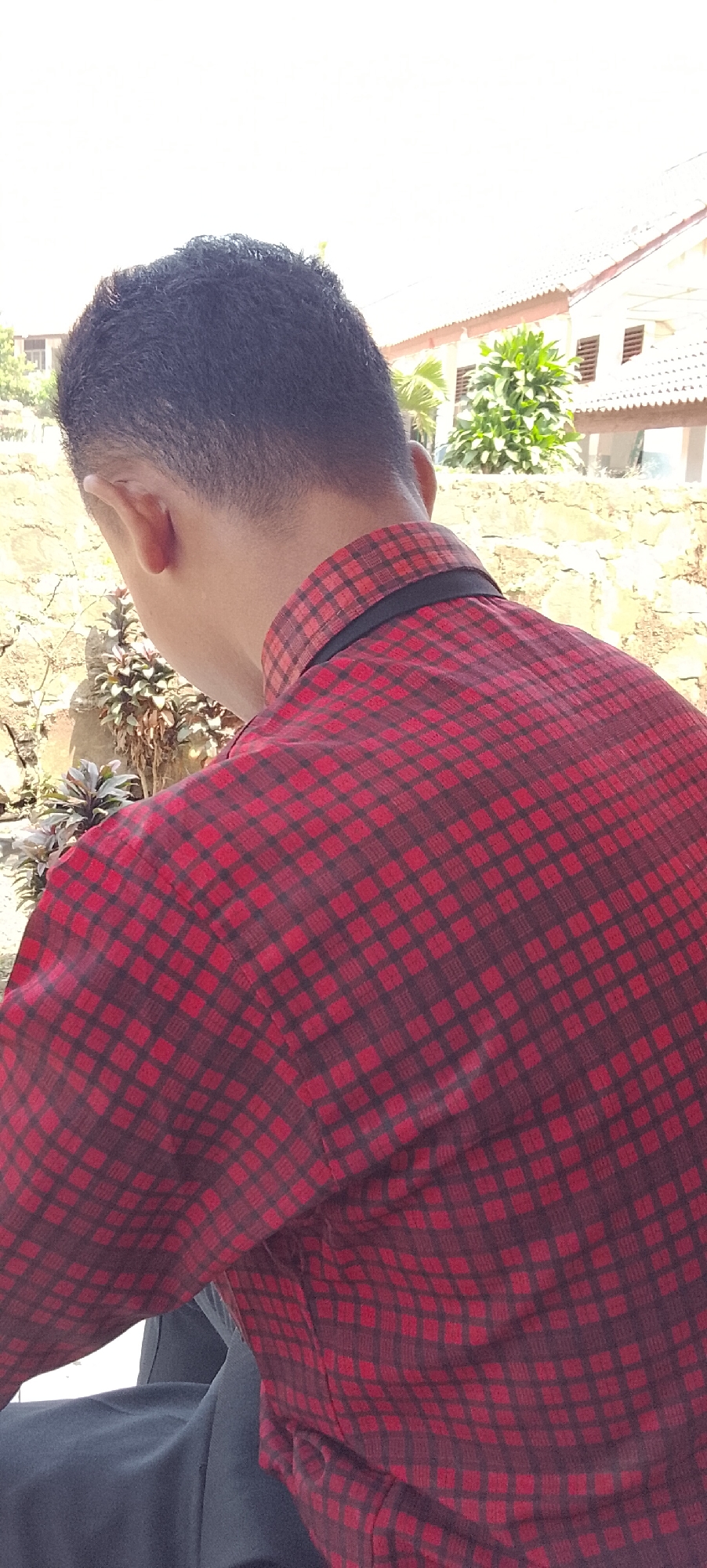 The image shows the back of a young man from the shoulders up, wearing a red and black checkered collared shirt. He is outdoors, with a blurred background of a building and some foliage. The man's hair is dark and cut short. A stone wall and some plants are visible to his left. The sky is bright and appears to be overcast. The scene is likely in a residential area in Tanjung Setia, Indonesia, given the style of the building and vegetation.