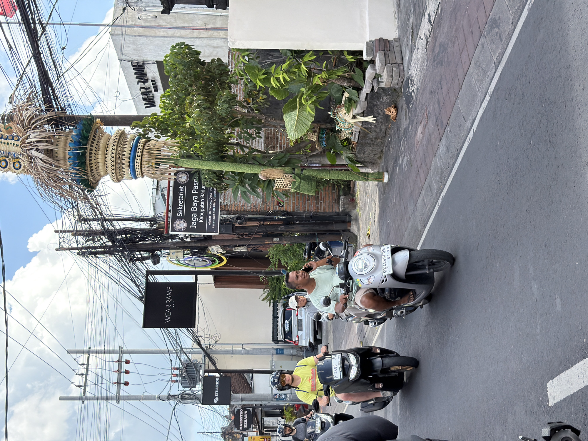 In an urban environment, a group of people are riding scooters on a paved road. The image captures a street scene with a variety of buildings, overhead electrical wires, and vegetation. The sky above is partly cloudy.
In the foreground, a person wearing a yellow t-shirt and sunglasses is riding a black scooter, looking forward. Next to them, a person in a light-colored t-shirt is riding a silver scooter and talking on a cell phone. Further back, another person is riding a black scooter. There are also glimpses of other vehicles and pedestrians.
The buildings lining the street display various signage, including "WEAR RAME" and "Sekretariat Jaga Baya Pasew". Decorative elements, possibly religious or cultural in nature, are visible near the roadside. The overall impression is a bustling street in what appears to be a tropical or sub-tropical location.