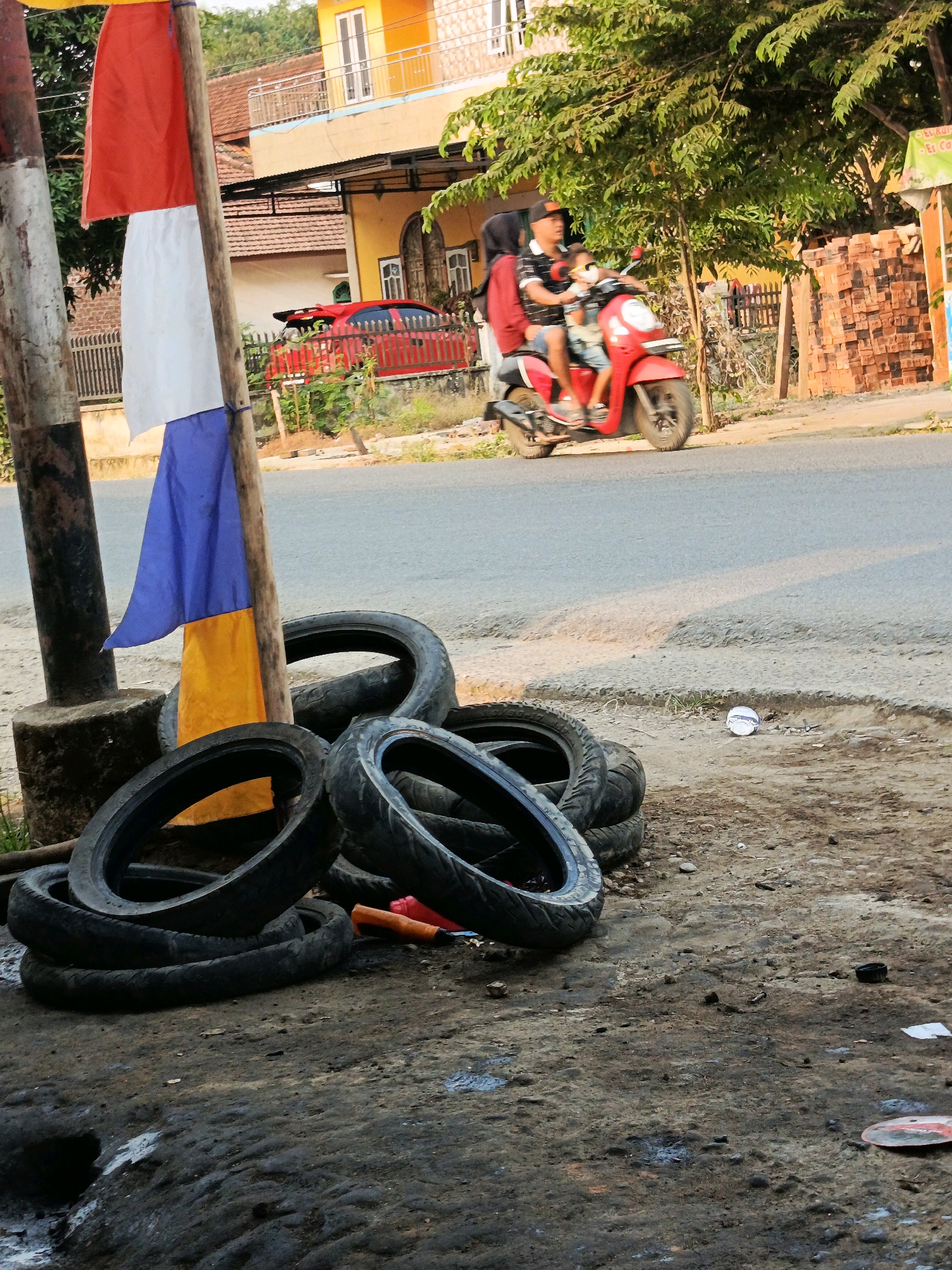The image captures a street scene in Megang Sakti I, Indonesia, focusing on a roadside area with various elements. In the foreground, a pile of discarded tires is visible on a rough, dirt surface. Behind the tires, a wooden pole stands, adorned with a tri-colored flag in red, white, and blue. The background reveals a street with a red vehicle driving on it, and beyond that, buildings with yellow and orange facades. A lush green tree with dense foliage provides shade to the right side of the frame. A family of three, a man, a woman, and a child, are riding a red motorcycle on the street. The man is driving, the woman is seated behind him, and the child is in front of her. The overall impression is a snapshot of everyday life in a semi-urban Indonesian setting, with a blend of commerce, transportation, and residential elements.