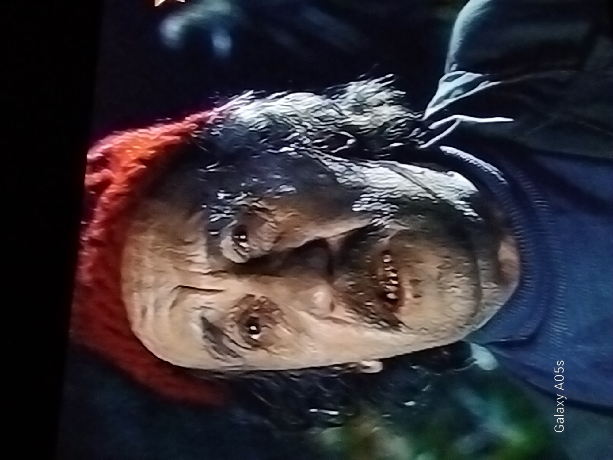 The image is a close-up, slightly grainy, portrait of an older man wearing a red knitted hat and a dark blue turtleneck sweater. His face is weathered and appears to be heavily made up to look aged or perhaps undead. He has dark, curly hair, a prominent mustache, and visible teeth that are discolored and uneven. His eyes are open and looking forward. The background is dark and out of focus, with some hints of green foliage. A watermark "Galaxy A05s" is visible in the bottom left corner, indicating the image was captured with a Samsung Galaxy A05s phone. There is no indication of the city of Druzhba, Russia, in the image.