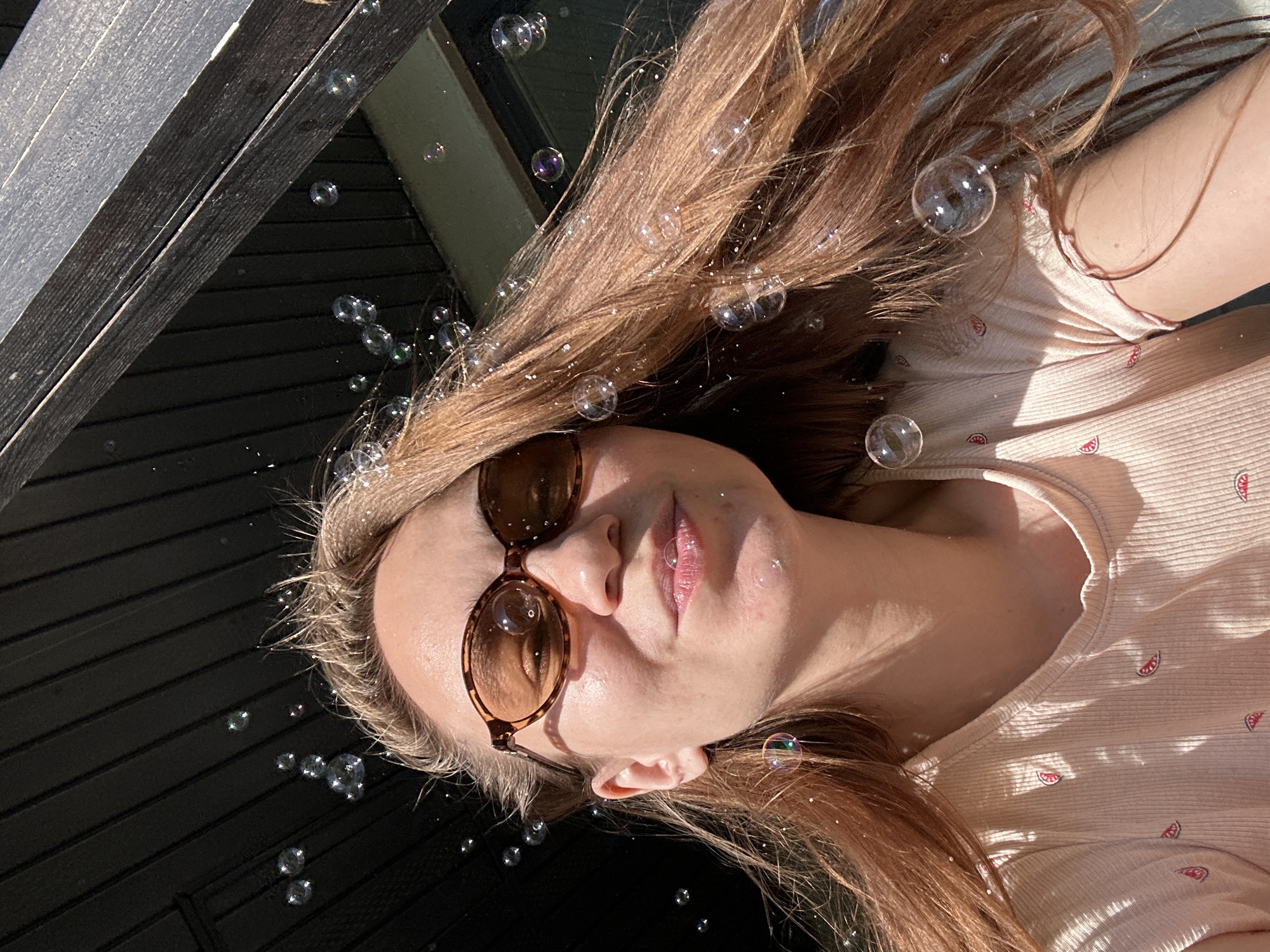 A woman with fair skin and light brown hair is lying down, wearing sunglasses. She is positioned in front of a dark wooden surface, likely a deck or similar structure. Bubbles are scattered around her and in her hair. She is wearing a light-colored shirt with a small watermelon pattern. The location is not discernible from the image.