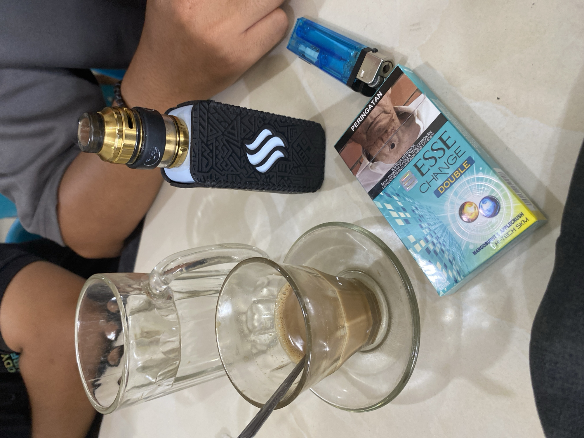 The image captures a scene in Lhokseumawe, Indonesia, featuring several items placed on a light-colored surface, possibly a table. Two people are partially visible in the frame.
The objects include a vaping device with a gold atomizer and a black protective sleeve, a blue lighter, a pack of "Esse Change Double" cigarettes, and two clear glass mugs, one of which contains a light brown liquid, likely coffee, with a spoon inside. The pack of cigarettes includes a warning label in Indonesian.