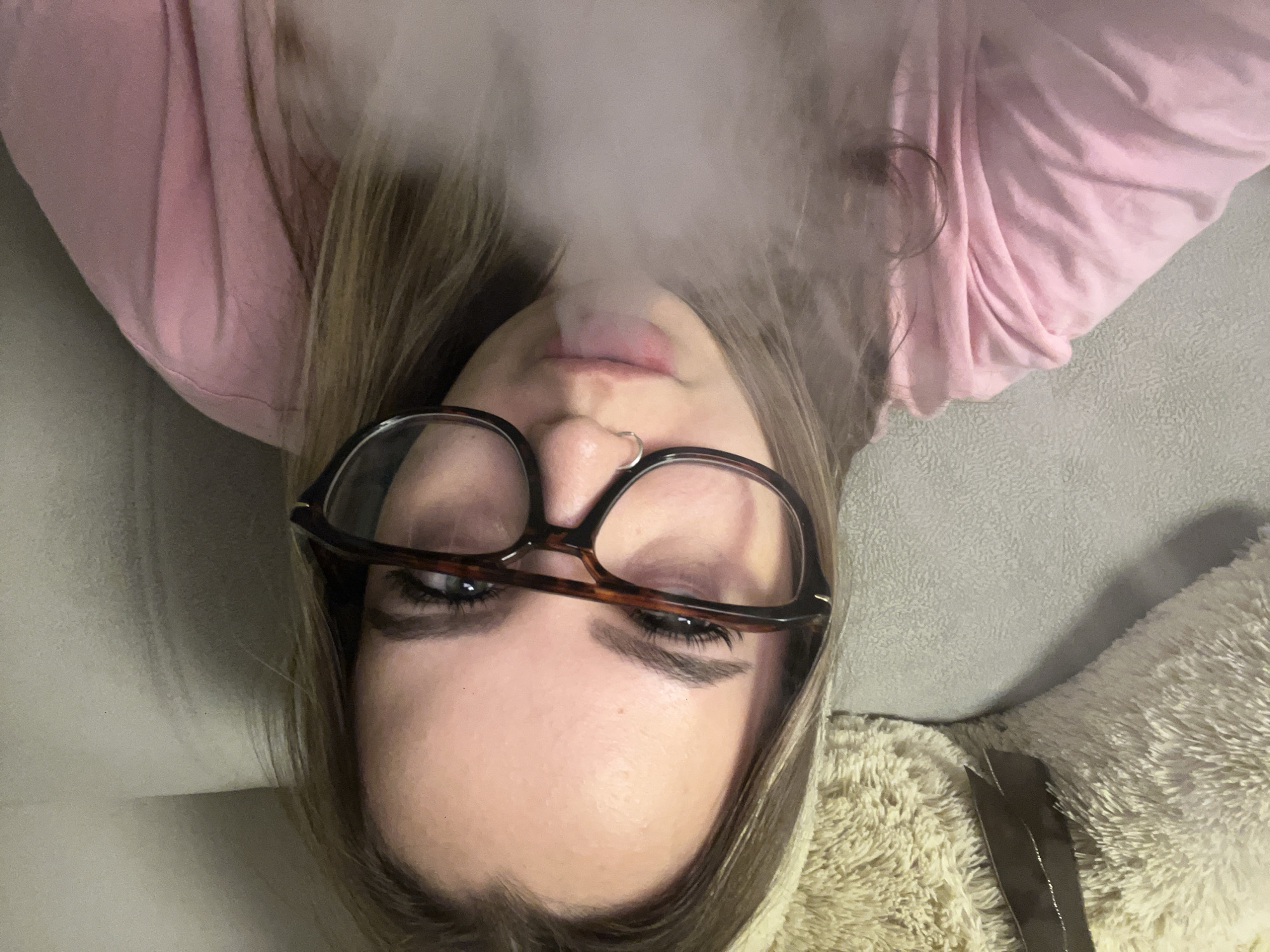 A young woman with blonde hair and brown-rimmed glasses is lying on a gray couch, wearing a pink long-sleeved shirt. She is exhaling smoke. A beige plush toy is visible to her right. The location cannot be determined from the image.