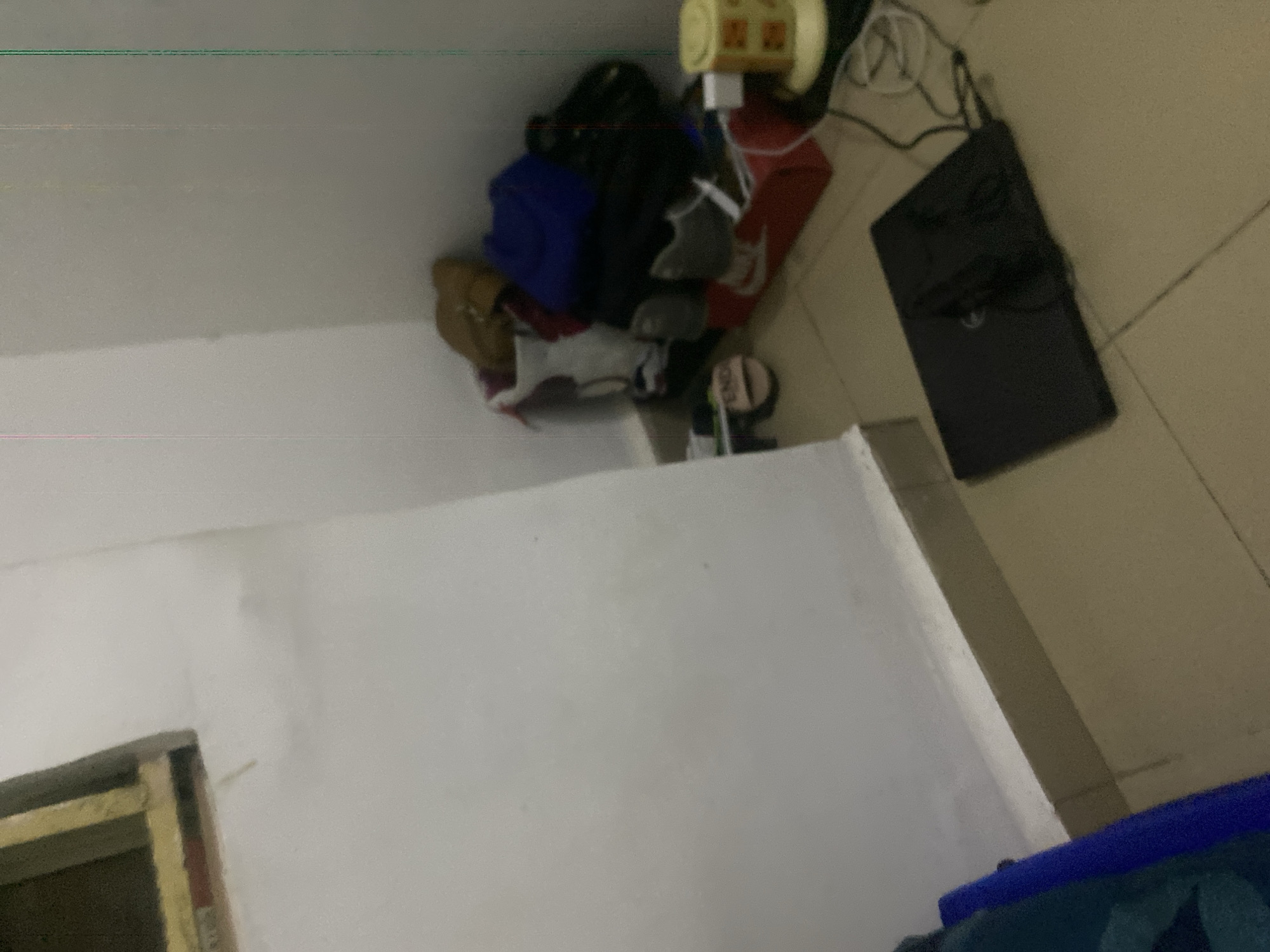 The image depicts an indoor scene, likely a room, with a corner where various items are piled up. In the foreground, a closed black laptop is placed on a tiled floor. To the right of the laptop, a yellow object with a grid pattern, possibly a small appliance or a charger, is visible. The corner area is cluttered with bags, shoes, and what appears to be clothing. Among the items in the corner are red shoes with white accents, a blue bag, and a brown bag. A white plastic bag is also present. A small, round, beige object with text that reads "END" is on the floor near the pile. The walls are painted white and beige, and there is a suggestion of a window frame in the upper left corner. The lighting is dim, and there is a slight blur to the overall image, potentially due to motion or low light conditions. The scene does not contain any individuals. Based on the provided context, this scene is located in Howayi, Nigeria.