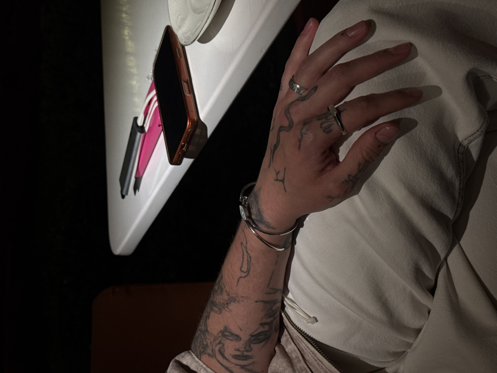 This low-light image provides a close-up view of a person's tattooed arm and hand, captured in an indoor setting, likely in Yerevan, Armenia. The dim ambient light suggests the scene takes place during the evening or night.
The hand, adorned with two silver-toned rings on different fingers, showcases a distinctive tattoo of a snake winding across its back. The forearm is extensively covered with various tattoos, including a detailed stylized face on the inner forearm and other abstract designs. A simple, polished silver-toned bracelet is worn on the wrist. The person is dressed in a light-colored garment, possibly a jacket or hoodie, visible by its textured fabric and a part of a zipper.
Resting on a white surface, likely a table, are several objects. A smartphone, encased in a copper or rose gold colored cover, is prominently placed. Beside the phone, several pens or markers in pink, black, and white are visible. A portion of a white ceramic plate is also seen in the upper left corner of the frame. The background is largely dark, emphasizing the focused subjects and contributing to the intimate, calm atmosphere of the moment. No discernible text is visible on any of the objects or tattoos.
