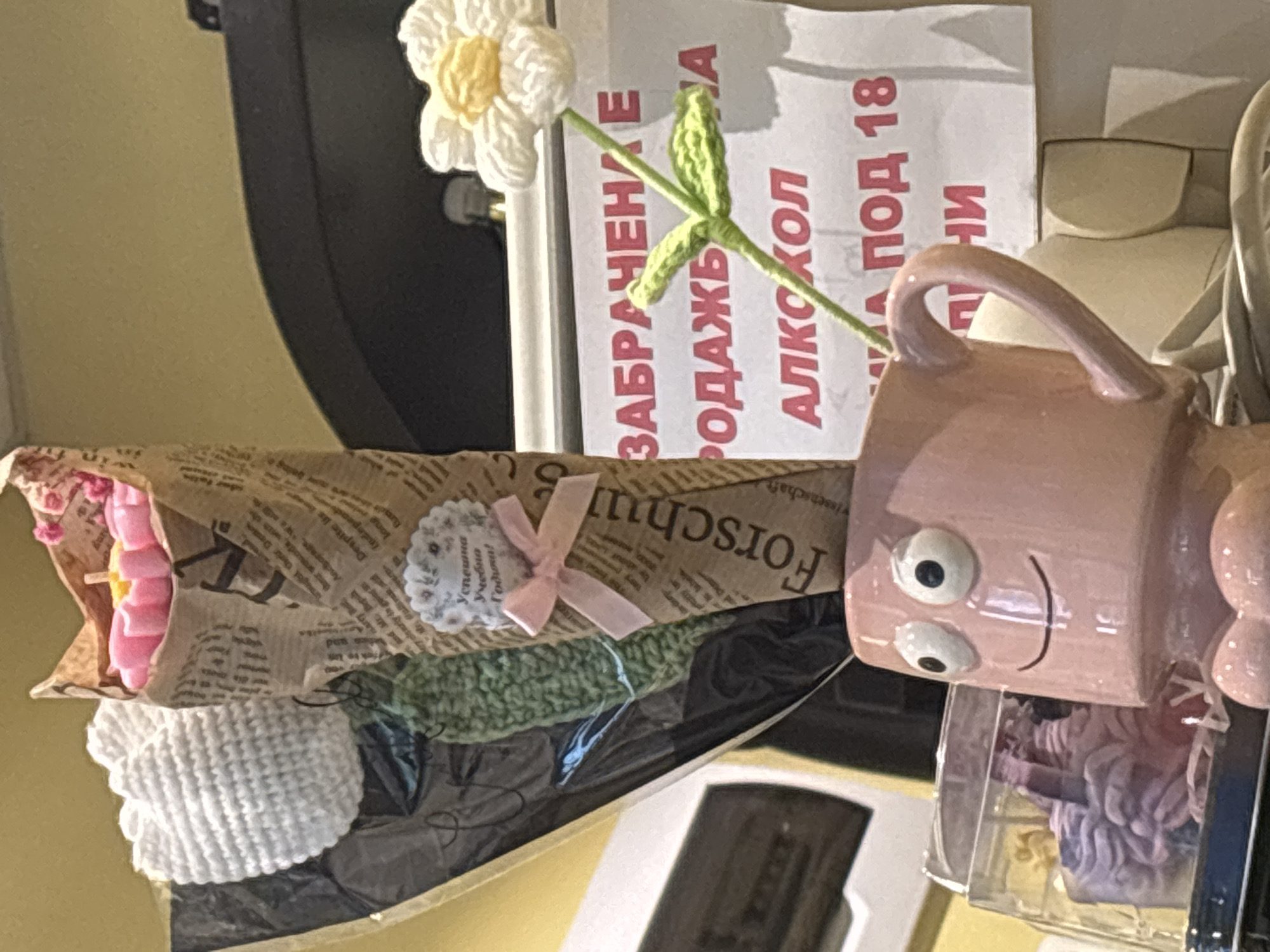 The image shows a collection of objects, likely arranged for display or sale. In the background, there's a sign in Bulgarian prohibiting the sale of alcohol to individuals under 18 years of age. In front of the sign is a crocheted white daisy. There is also a bouquet wrapped in newspaper-like wrapping, featuring a tag with "Успешна Учебна Годом!" printed on it, tied with a light pink bow. Next to the bouquet is a pink mug with googly eyes. The setting appears to be indoors. There is insufficient information available to confirm the location is in Burgas, Bulgaria.