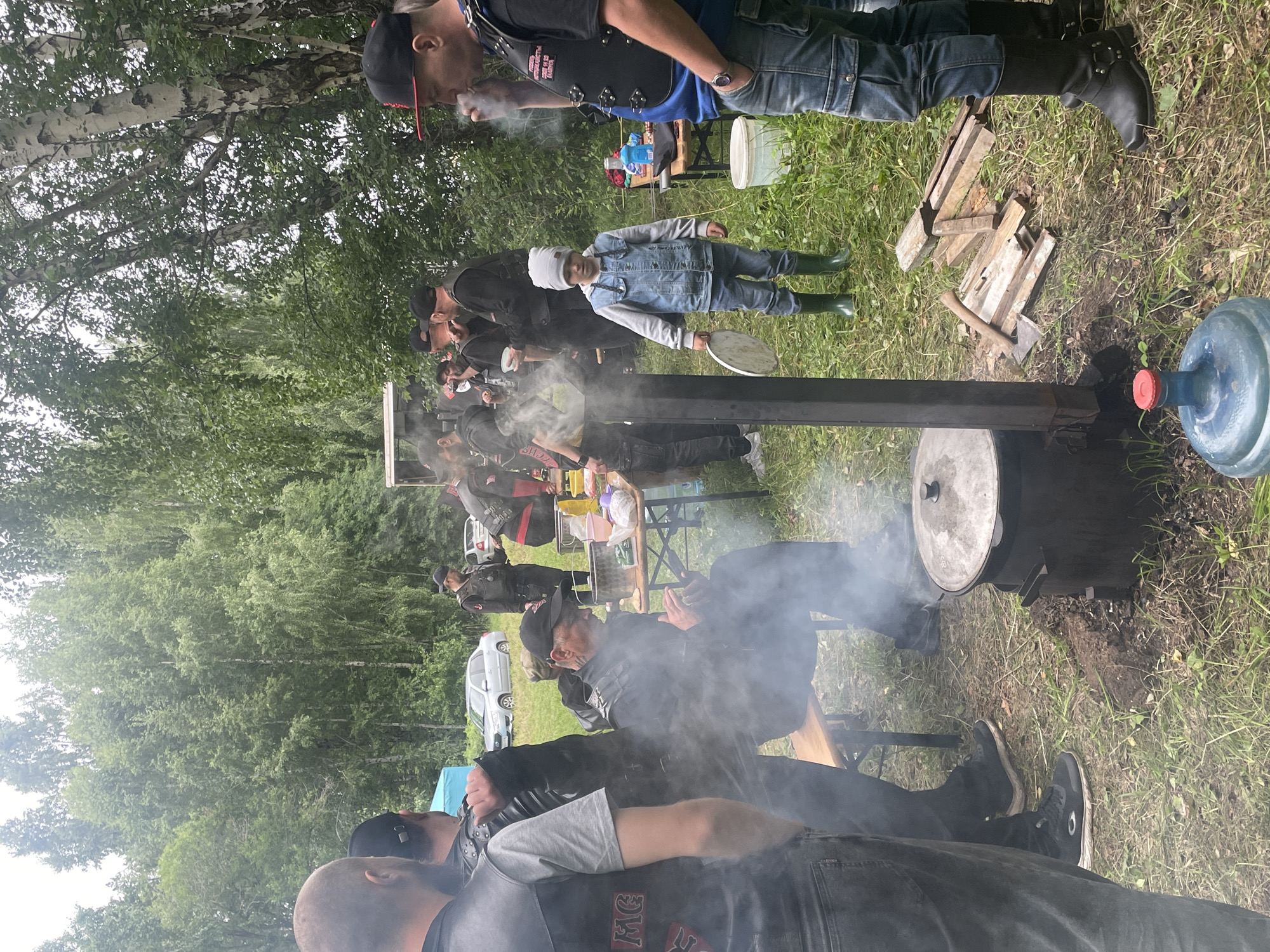 A group of individuals, some wearing biker vests, are gathered outdoors in what appears to be a wooded area near Il'ichevskii, Russia. A large pot is being heated over an open fire, with smoke rising from it. A young child in a denim jacket and green boots stands nearby, holding a plate. A table with food and drinks is set up to the right, with several people interacting around it. The ground is covered in grass and fallen leaves, and trees form a dense background. Several cars are visible in the distance.