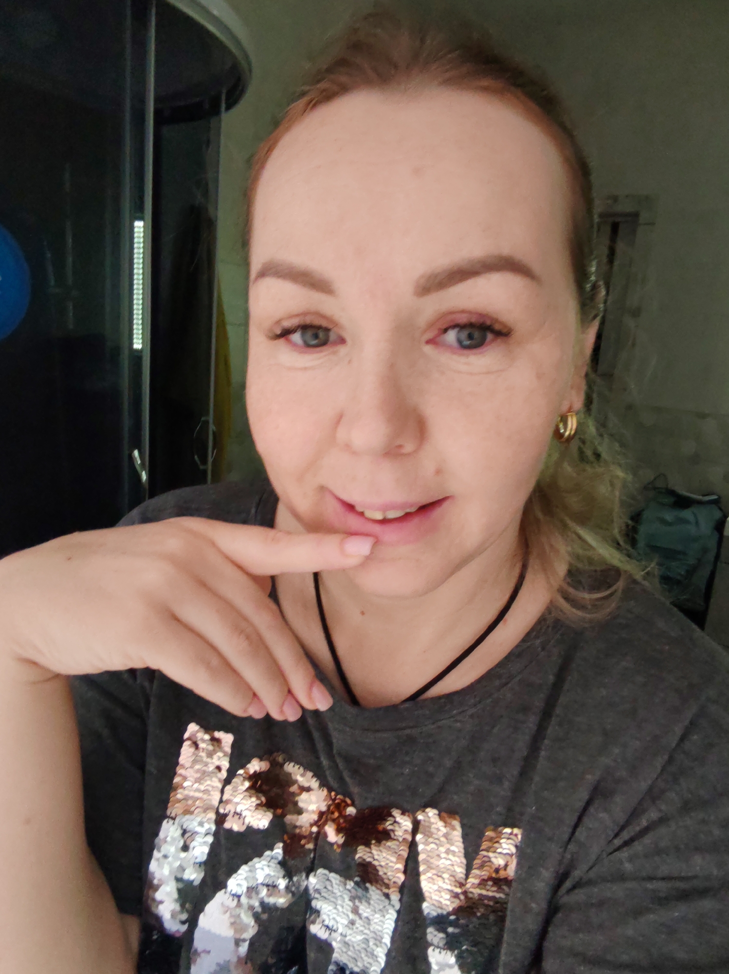 A selfie of a woman with blonde hair in a ponytail wearing a gray t-shirt with sequin letters, gold hoop earrings, and a black cord necklace. She is holding her index finger to her lower lip. Behind her is a dark shower stall and a window. The photo was likely taken indoors. It is not possible to verify the location as Chaikovsky, Russia, with the provided image.