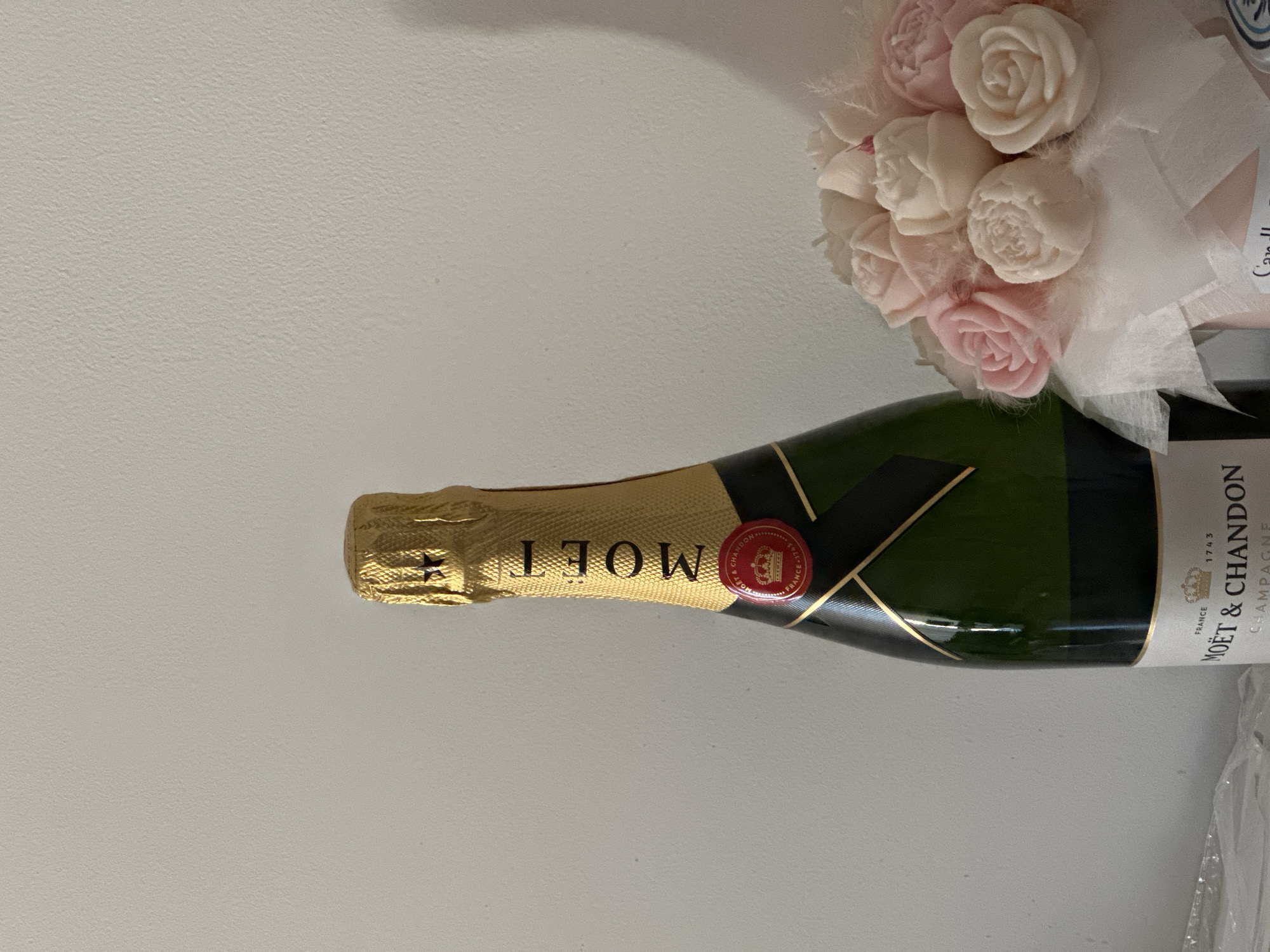 The image shows a bottle of Moët & Chandon champagne lying horizontally on a white surface, next to a bouquet of light pink and white roses wrapped in white tulle. The champagne bottle has a gold foil top and a black label with gold lettering. The roses appear to be made of soap or wax. The setting cannot be determined to be in Budva, Montenegro from the image alone.