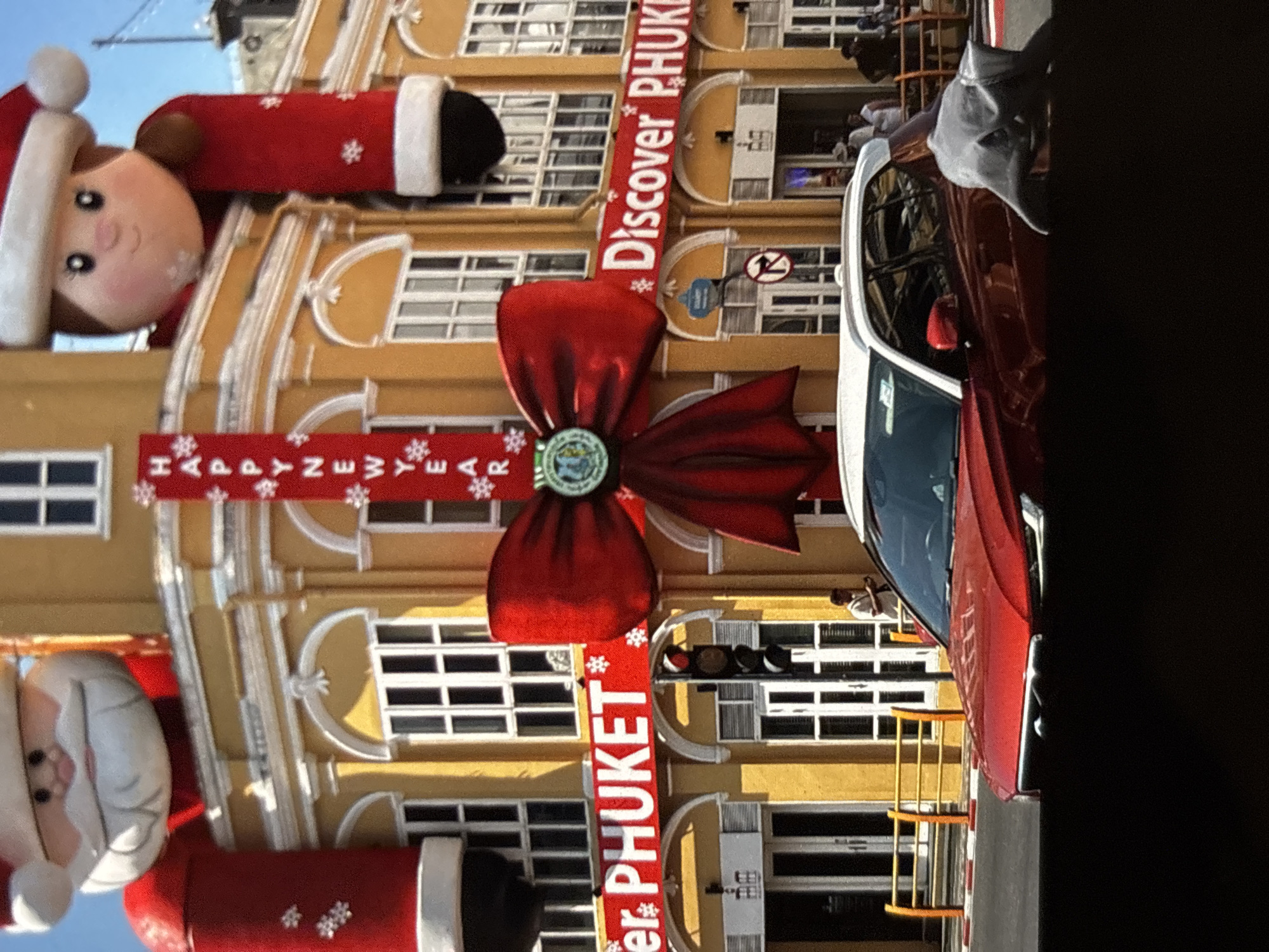 The image captures a festive street scene in Chalong, Thailand, decorated for the New Year. Two large inflatable figures, resembling Santa Claus and a child with Santa hats, are prominently displayed on the facade of a yellow building. A vertical red banner with white lettering spells out "HAPPY NEW YEAR" with snowflake embellishments. A large red bow is affixed to the building, and two horizontal red banners with white lettering read "er. PHUKET" and "Discover PHUKET". In the foreground, a red SUV is parked on the street, partially obscuring a grey inflatable dolphin. Several people are visible on the sidewalk and entering the building. The environment suggests a commercial area with shops and pedestrian activity.