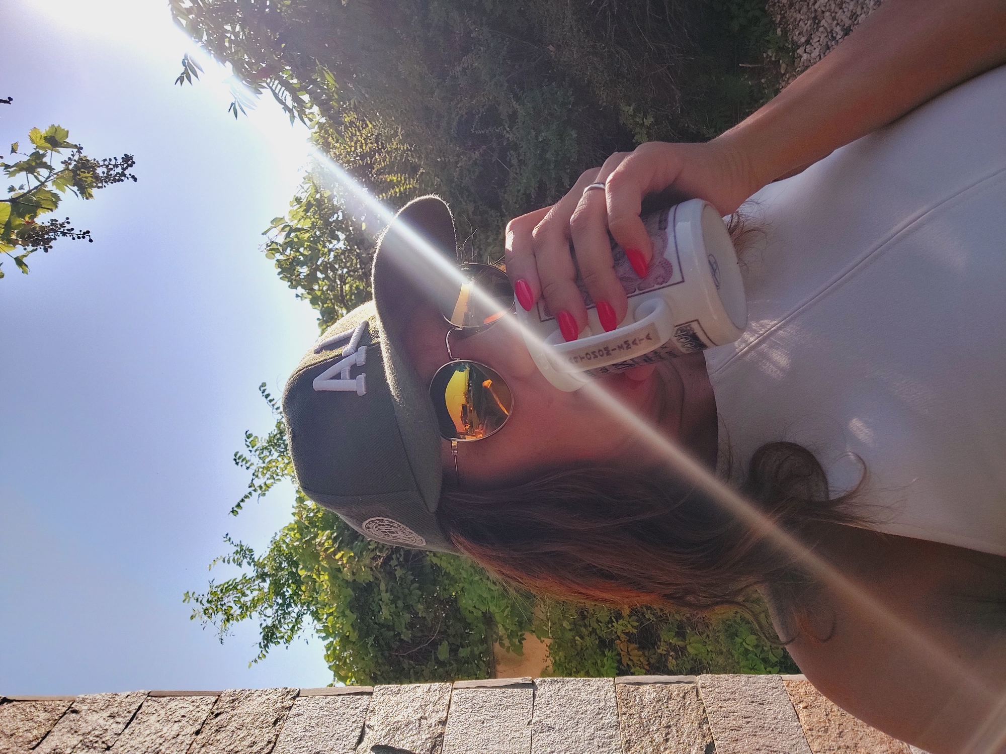 A woman wearing a green baseball cap and sunglasses is drinking from a white mug in an outdoor setting. She has long brown hair and red fingernails. The sun is shining brightly, casting a lens flare across the image. In the background, there are green trees and a stone wall. The woman is wearing a white top. The mug has a pattern on it.