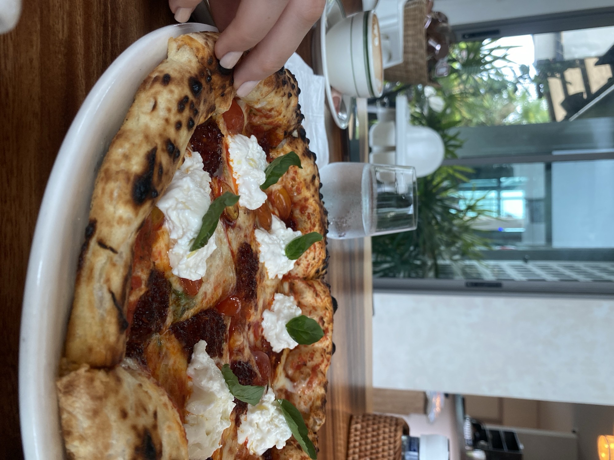 A photograph shows a partially eaten pizza on a white plate with a raised edge, likely at a restaurant in Choeng Thale, Thailand. The pizza is topped with tomato sauce, fresh mozzarella cheese, cherry tomatoes, and basil leaves. The crust is puffy and blistered, indicating it was baked in a hot oven. A hand is reaching for the pizza. Next to the plate is a glass of water and a cup of coffee. The background features a blurred view of greenery and a window, suggesting an outdoor or semi-outdoor dining setting.