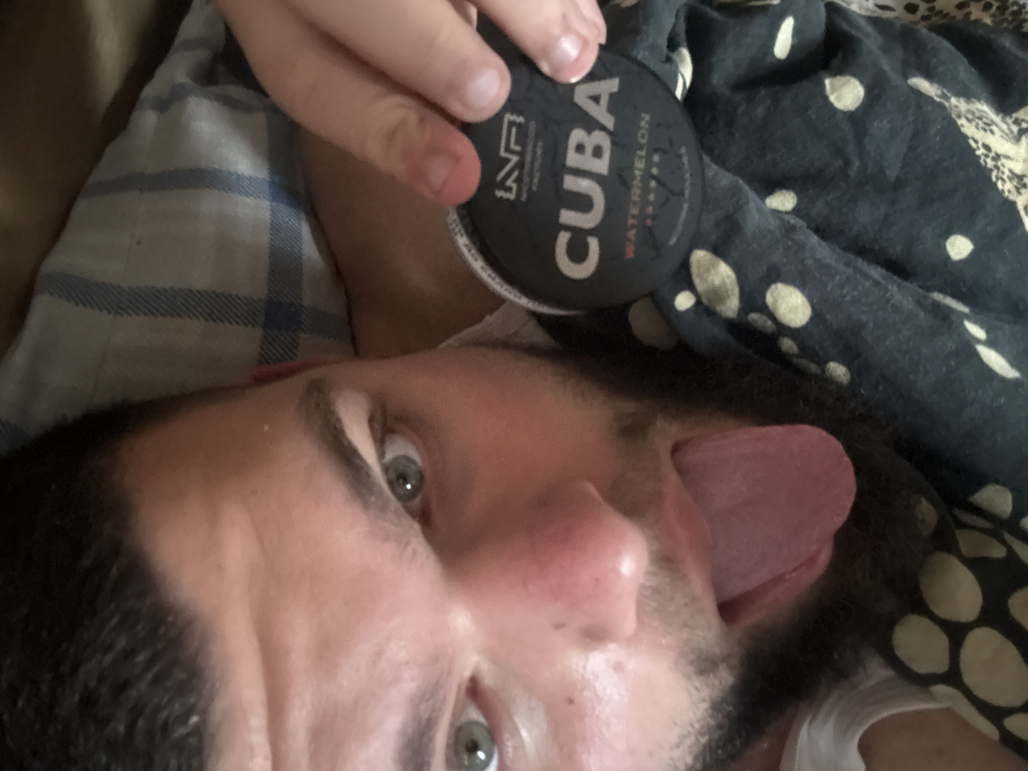 A man lies on patterned bedding with his tongue extended. He is holding a black tin of "CUBA Watermelon" nicotine pouches. The location is unknown and there is no information in the image to place it in Burgas, Bulgaria.