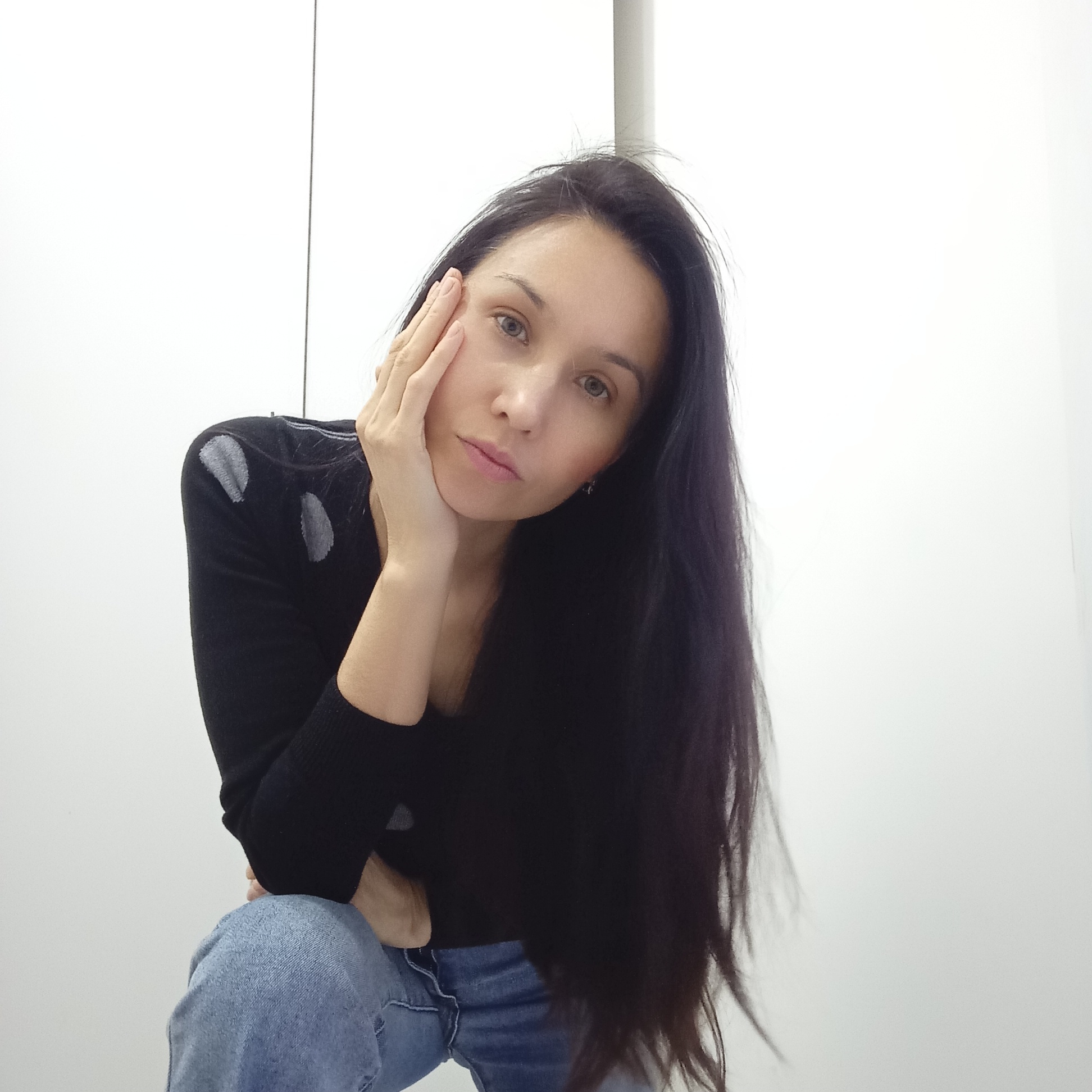 A woman with long, dark hair is posing indoors. She is wearing a black long-sleeved shirt with gray patterned accents on the shoulders, and blue jeans. She is sitting with one hand resting on her cheek and the other arm resting on her knee. The background is a plain, white wall. The image doesn't depict any distinctive characteristics or elements that allow to pinpoint a specific location in the city of Izhevsk, Russia.