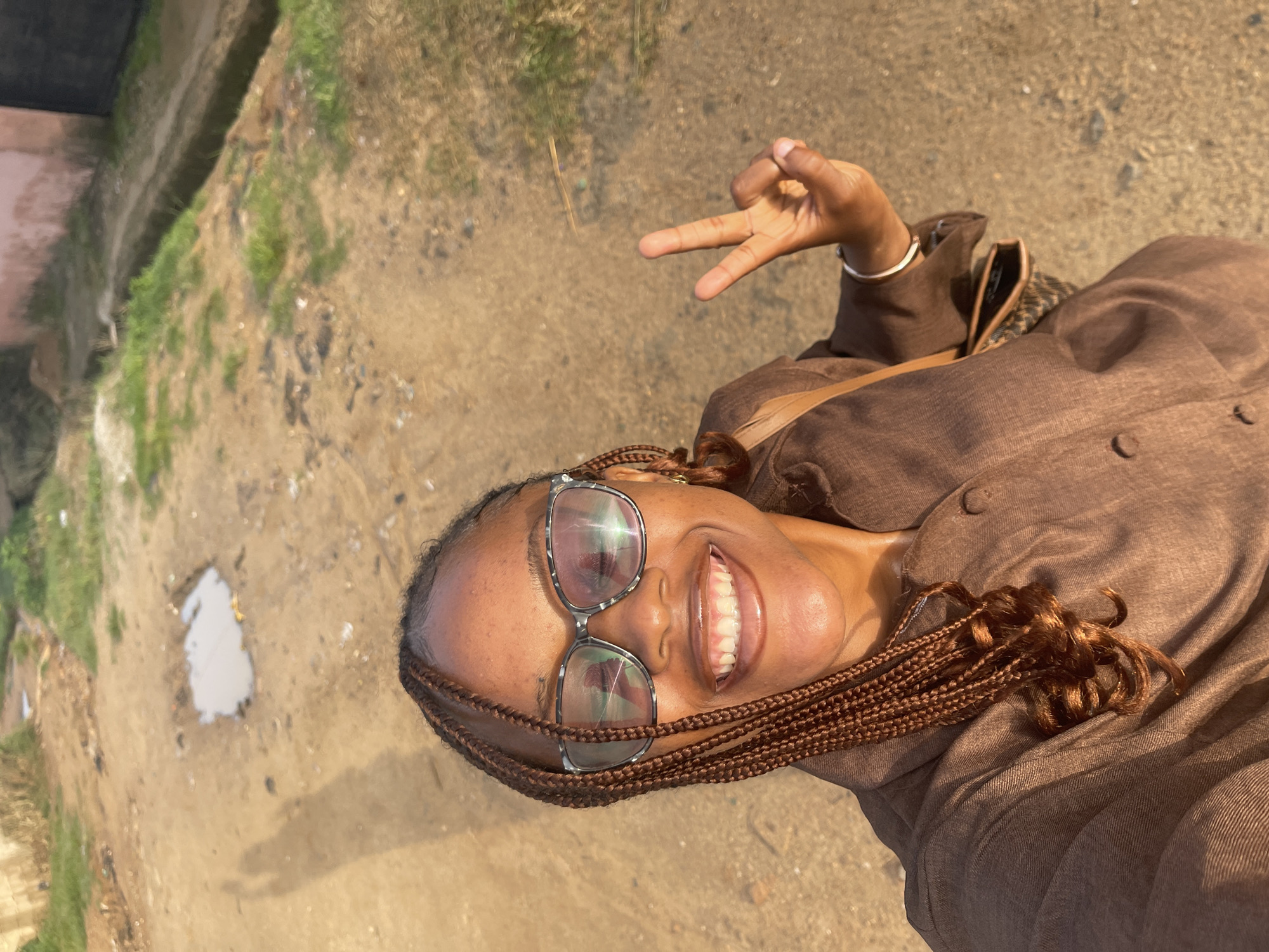 A young Black woman with braided hair and glasses is taking a selfie outdoors. She is smiling and holding up two fingers in a peace sign. She is wearing a brown collared shirt. In the background, there is a dirt path with some puddles and overgrown grass. To the right, there is a concrete ditch and a pink-ish wall with a dark metal gate. The scene appears to be in Rafin Par, Nigeria, given the context.