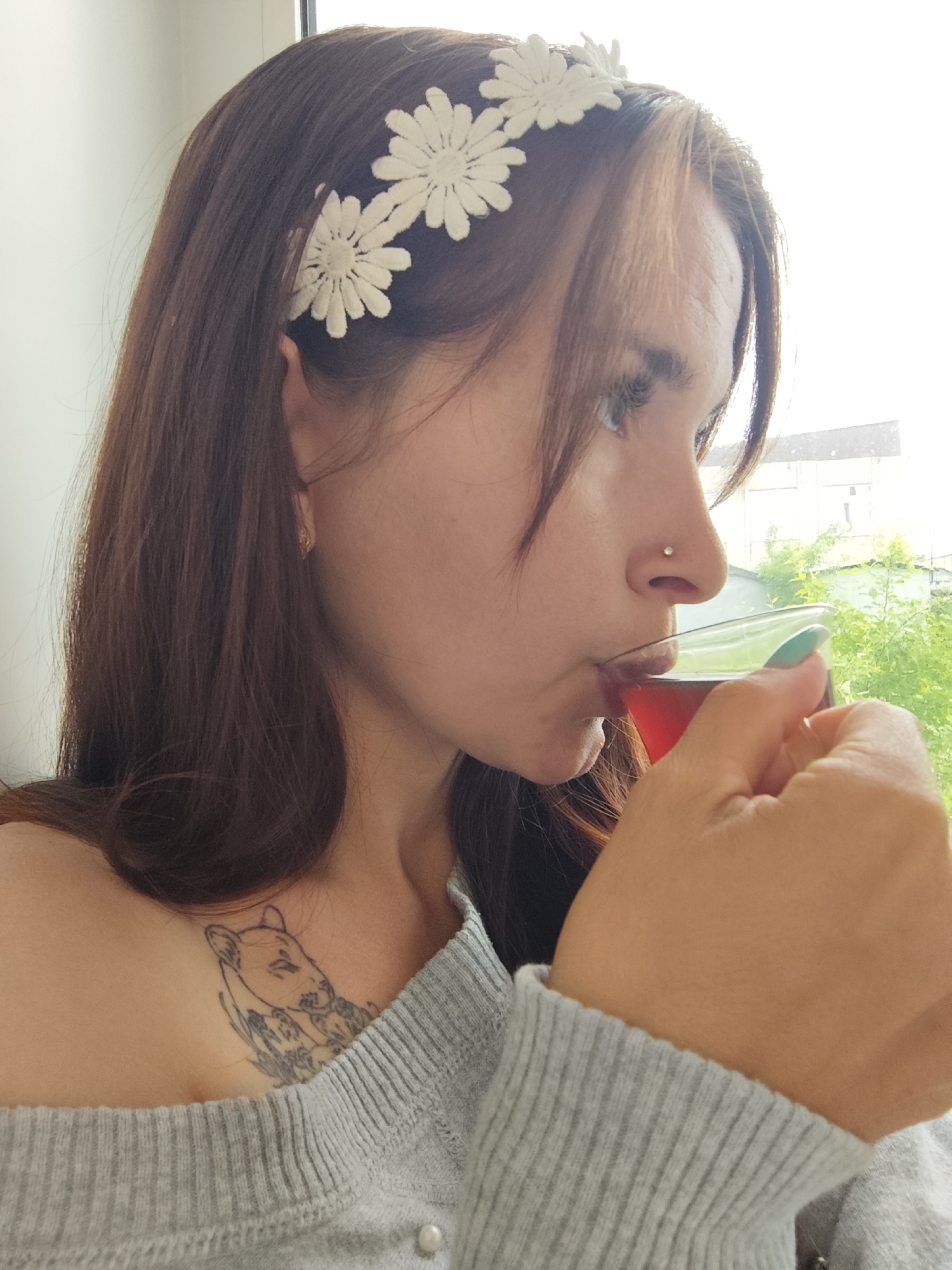 A woman with dark hair and a daisy-chain headband is drinking from a glass of red liquid. She has a nose piercing and a tattoo of a lion or similar feline on her upper chest. She is wearing a grey off-the-shoulder sweater. The background shows a window with foliage and a building visible through the glass. The location is Фоки, Russia, as the prompt indicates.