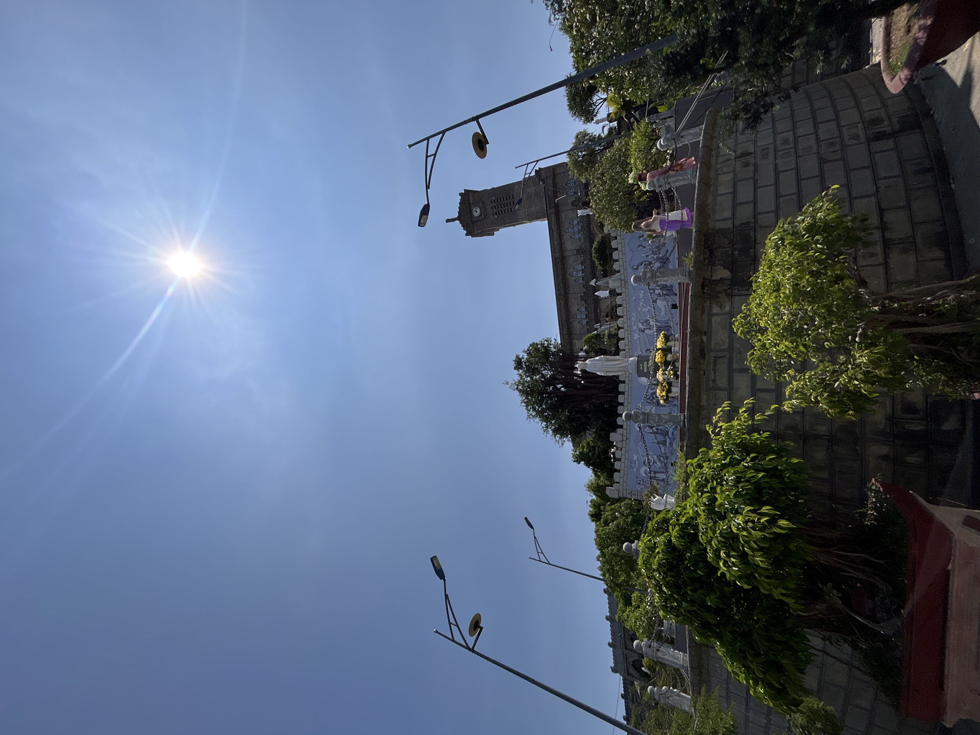The image depicts a low-angle view of the Holy Mary Mother Church in Nha Trang, Vietnam. The church features a tall, rectangular clock tower visible against a bright blue sky with the sun shining intensely. Below the church, there is a raised platform containing religious statues and images behind a low castle-like wall. Lampposts line the foreground. Trees and foliage surround the church and platform. Two individuals, presumably tourists, stand on the wall next to the church. The environment appears to be a sunny day.