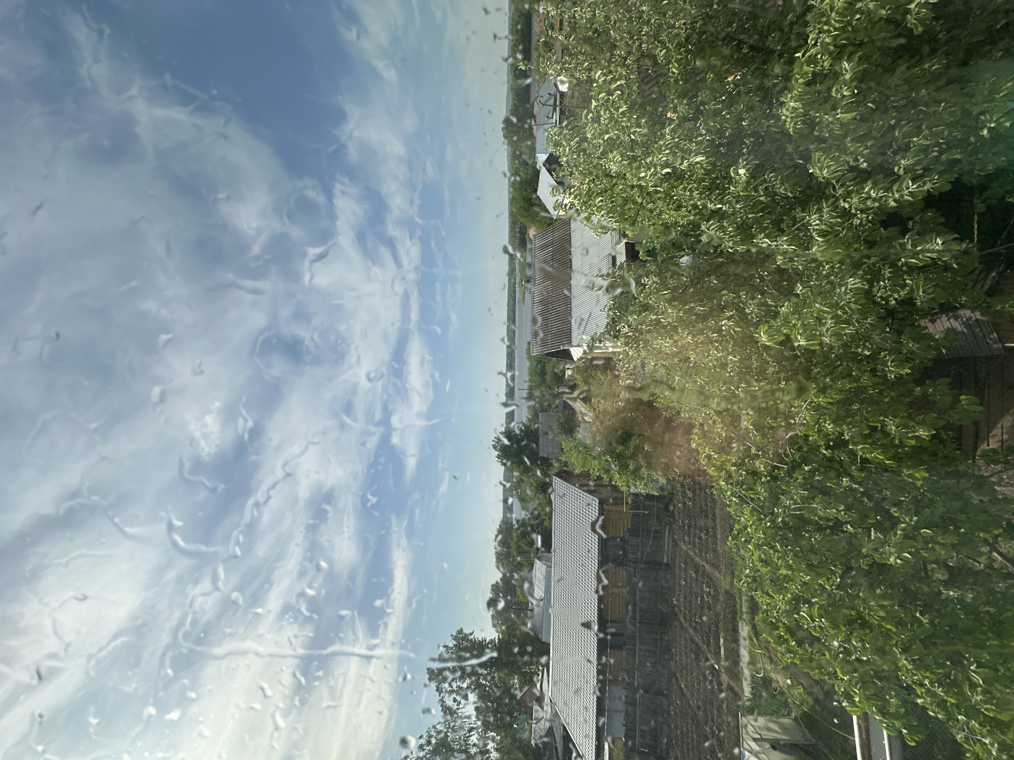 The image, taken from indoors looking out through a wet window, shows a view of a residential area in Chaikovsky, Russia. The foreground of the image, affected by water droplets on the window, showcases a cloudy sky with streaks of blue. Beyond the window, there is a cluster of houses with grey-colored metal roofs. Lush green trees are seen interspersed throughout the scene, especially to the right side of the frame, partially obscuring some buildings. The location is likely a residential neighborhood with a mix of houses and trees under a partly cloudy sky.