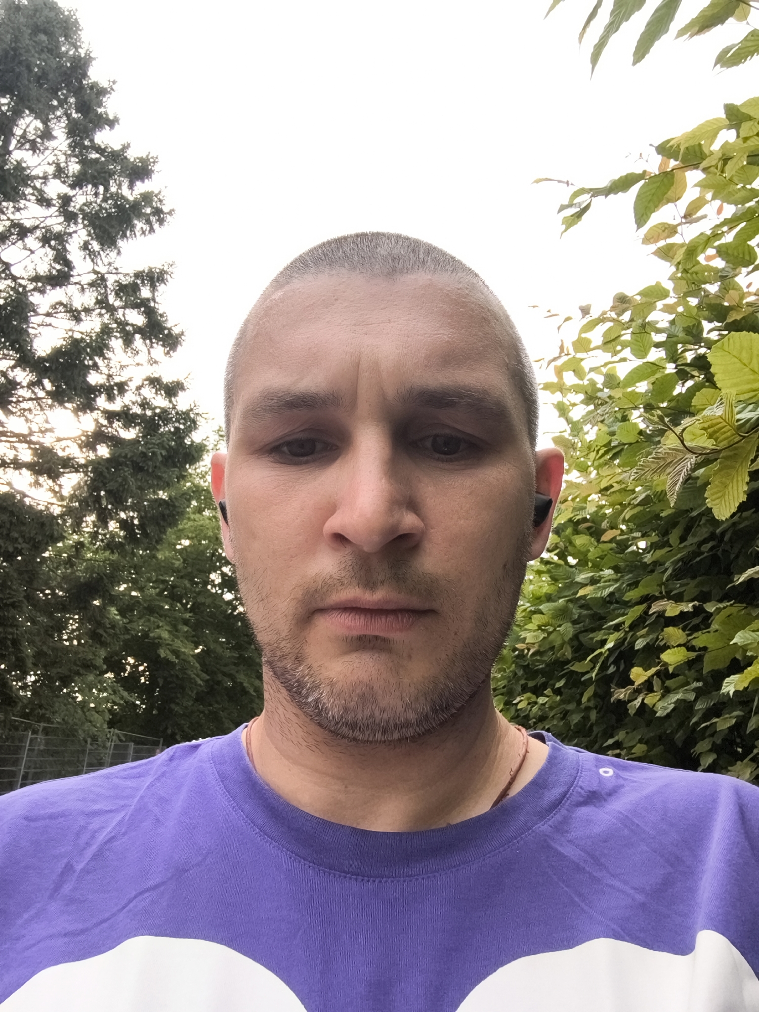 This is a selfie taken outdoors, close-up on a person's face and upper chest. The subject is a light-skinned male with a shaved head and stubble on his face. He is wearing a purple t-shirt with a large white design on it. He has dark eyes and appears to be looking directly at the camera. He is wearing a black wireless earbud in his right ear.
The background is blurred with trees on either side of the subject. On the left, there are dark green evergreen trees and on the right, there are trees with lighter green leaves. The sky is visible at the top of the image, appearing overcast. There is a wire fence visible behind the trees on the left side of the image.
Based on the available information, the image was likely taken in Wolgast, Germany, given the visual context of outdoor foliage and an overcast sky, which is common for the region.