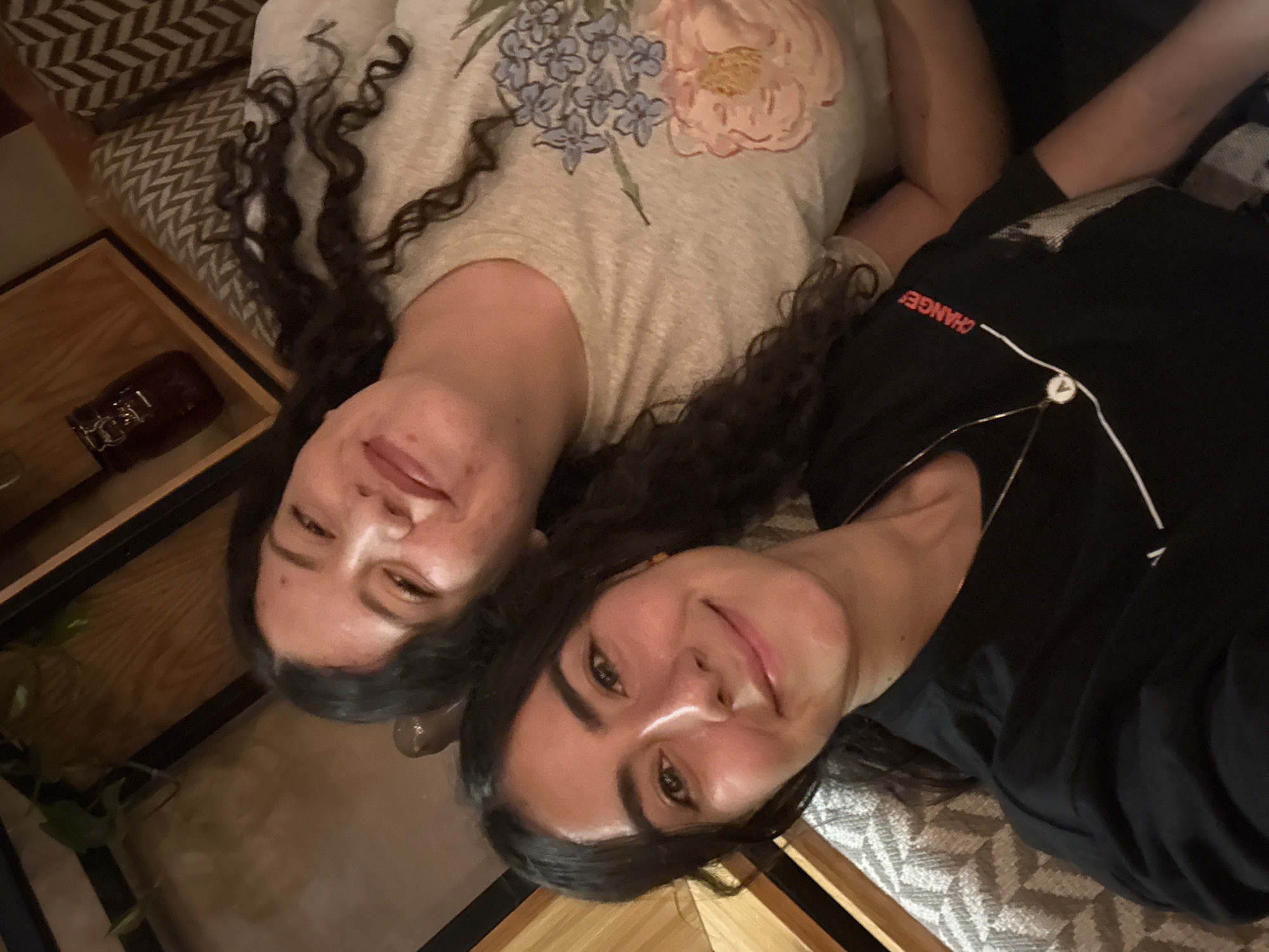 Two women lie side-by-side on a patterned cushion, likely indoors in Puebla, Mexico. The woman on the left has dark curly hair and is wearing a light-colored t-shirt with a floral design. The woman on the right has dark, straight hair and is wearing a black t-shirt with the word "CHANGE" printed on it. Both women are looking at the camera and smiling. The environment appears to be a seating area with wooden accents and some foliage in the background. A brown container can be seen on a wooden shelf next to them.