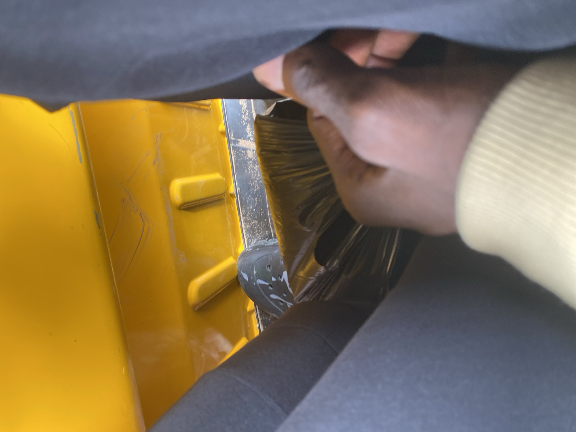 The image shows a close-up view of a person's hand reaching into a yellow object. The hand is dark-skinned and wearing a light-colored sleeve. The yellow object appears to be made of plastic and has some scratches on its surface. Inside the yellow object, there is a black, crinkled material that might be a trash bag. A green shoe with white splatters is also visible, partially obscured by the black material. The background is dark and blurry, suggesting an indoor setting or poor lighting conditions. The image is taken from a low angle, looking upwards. There is no visible text in the image. The context provided indicates the location is Jalingo, Nigeria.