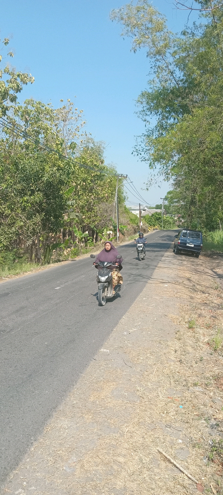 A vertical, eye-level, medium shot captures a bright, sunny day on a rural road in Kedukbembem, Indonesia. The asphalt road is bordered on the left by lush green trees and vegetation, and on the right by dry, straw-covered ground and more trees. A dark blue van is parked on the right side of the road. In the foreground, a person wearing a purple head covering and a patterned skirt rides a black motorcycle, traveling away from the viewer. Behind them, another person on a smaller black motorcycle is also moving away from the camera. The sky is a clear, light blue. Power lines stretch across the upper portion of the image.