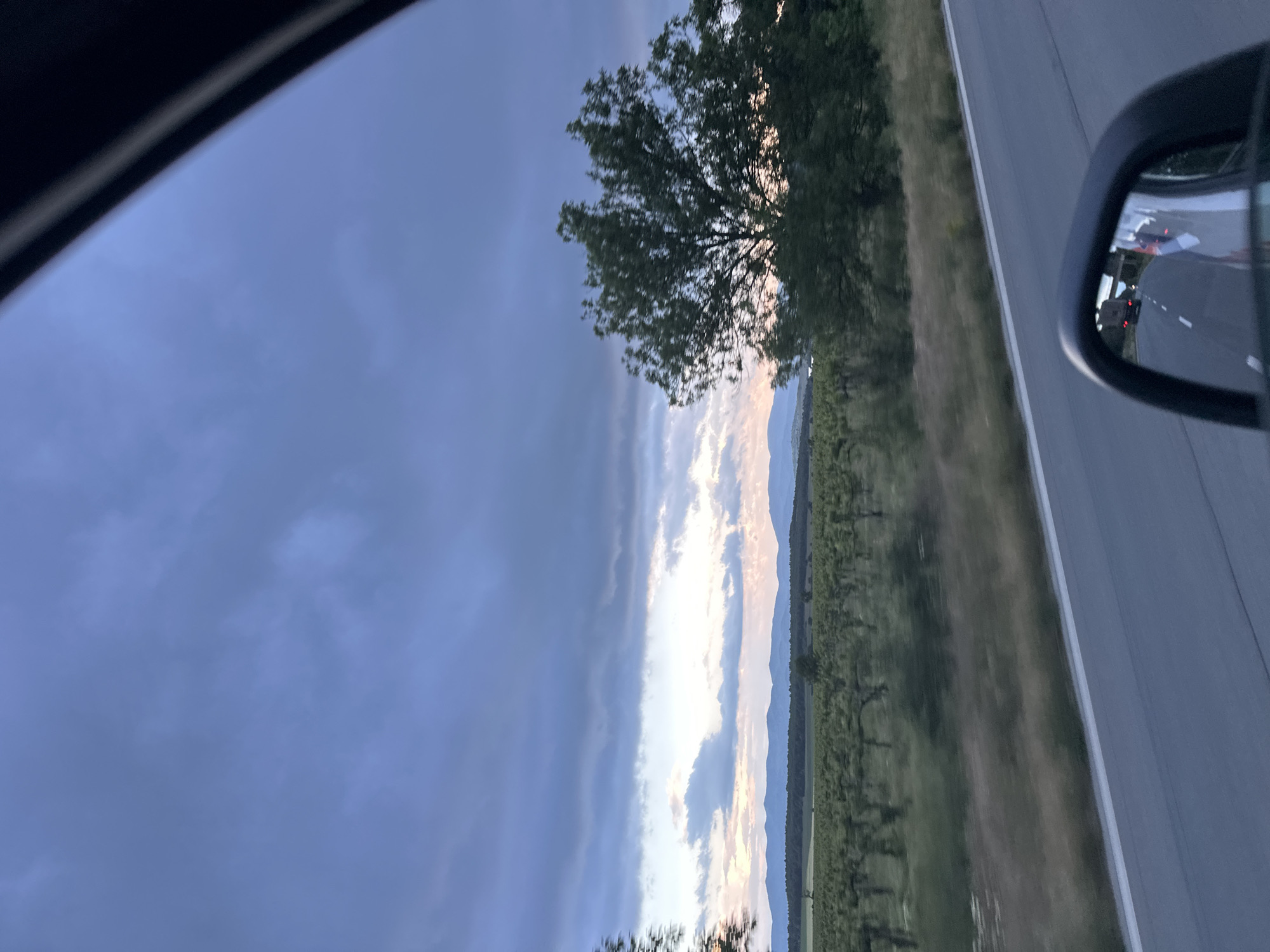A roadside view taken from within a car; the outside rearview mirror is visible in the upper right, reflecting other cars on the road. A gray, asphalt road runs along the right side of the frame. To the left of the road is a grassy median, beyond which lies a vineyard. In the distance, silhouetted mountains are visible underneath a band of light breaking through the clouds. The left side of the frame is dominated by a dark, cloudy sky. A tree is seen between the vineyards and the sky. The location is likely near Konare, Bulgaria based on the prompt.