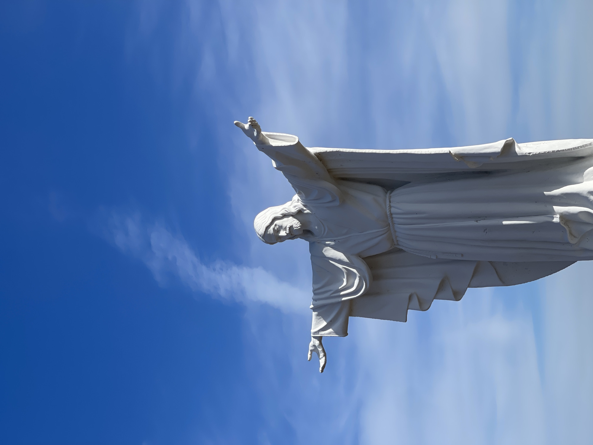 The image shows a low-angle view of the statue of Jesus Christ in Krasnoyarsk, Russia, against a clear blue sky with sparse cloud cover. The statue is white, depicted with outstretched arms. The image is rotated counterclockwise approximately 90 degrees.