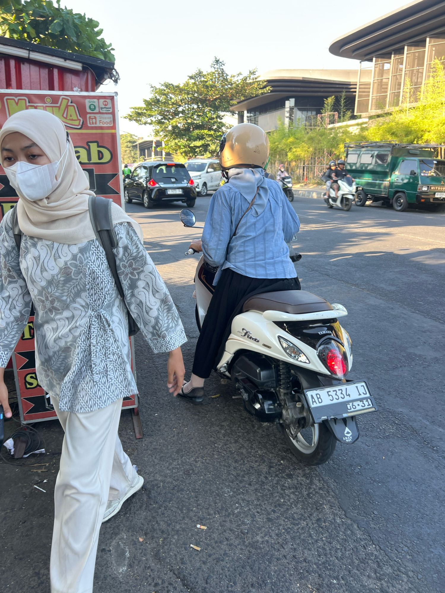 In Depok, Indonesia, on a paved road under a clear sky, a woman wearing a light-colored hijab, white face mask, and a patterned grey and white tunic over light-colored pants walks towards the left of the frame. She is carrying a dark backpack. To her right, a person in a blue long-sleeved shirt and black pants, wearing a light-colored helmet, rides a white Yamaha Fino scooter with a brown seat. The scooter's license plate reads "AB 5334 IJ" and "06-31". In the background, a black car, a white car, a green truck, and other vehicles are visible on the road. Trees and modern buildings with curved roofs are also present in the urban environment. A food stall with red and white signage indicating "Grab" and "Shopee Food" is partially visible on the left. Cigarette butts are scattered on the road surface.