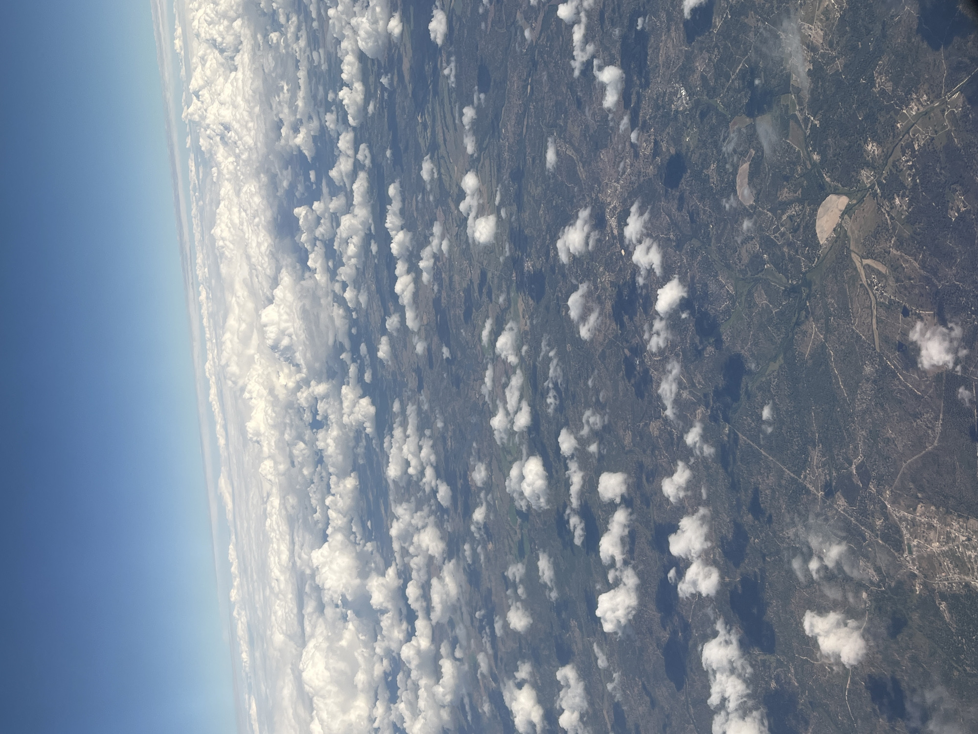 The image shows an aerial view of a landscape, with a focus on the transition between the sky/atmosphere and the terrain below. The sky is visible in the top left of the frame, showcasing a clear gradient from deep blue to a lighter, hazier blue near the horizon. Scattered, puffy white clouds partially obscure the view of the ground below, creating a dappled pattern of light and shadow.
The terrain appears to be rural, with a mixture of green vegetation, possibly forests or fields, interspersed with what seem to be bare patches or cultivated areas. The patterns suggest varying land use and natural features, potentially including small water bodies or drainage systems.
While there is no specific reference to the city of Coruche, Portugal within the image, the landscape type is representative of the general area. The image does not have any people, so there are no interactions or actions. It also does not show any objects or subjects except for the features listed.