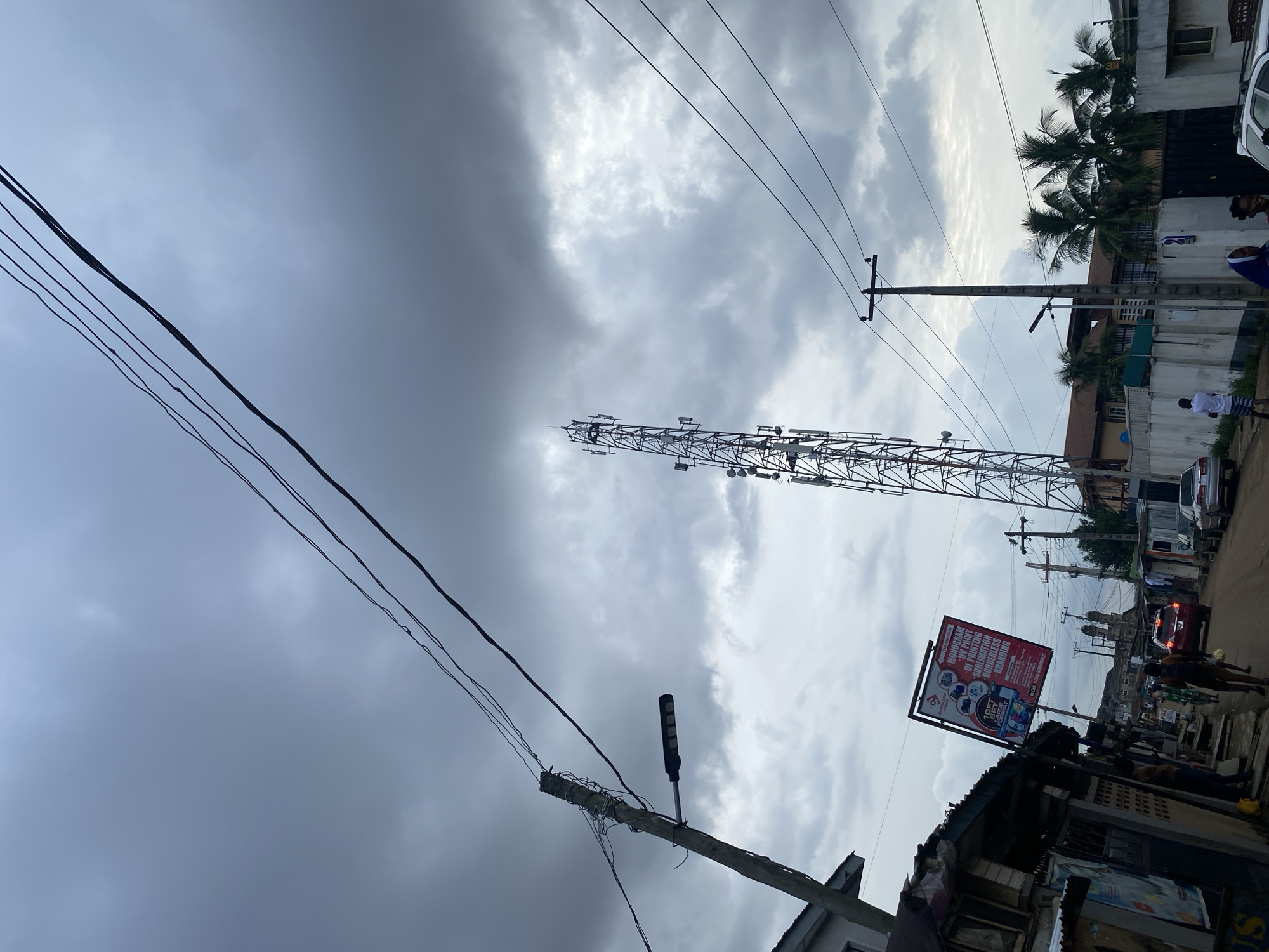The image depicts an urban scene, possibly in Lagos, Nigeria, characterized by overhead power lines and a telecommunications tower rising into a cloudy sky. A billboard advertising printing services is visible on the right side of the frame, positioned atop a structure. A street scene with buildings, vehicles, and pedestrians is discernible in the background.