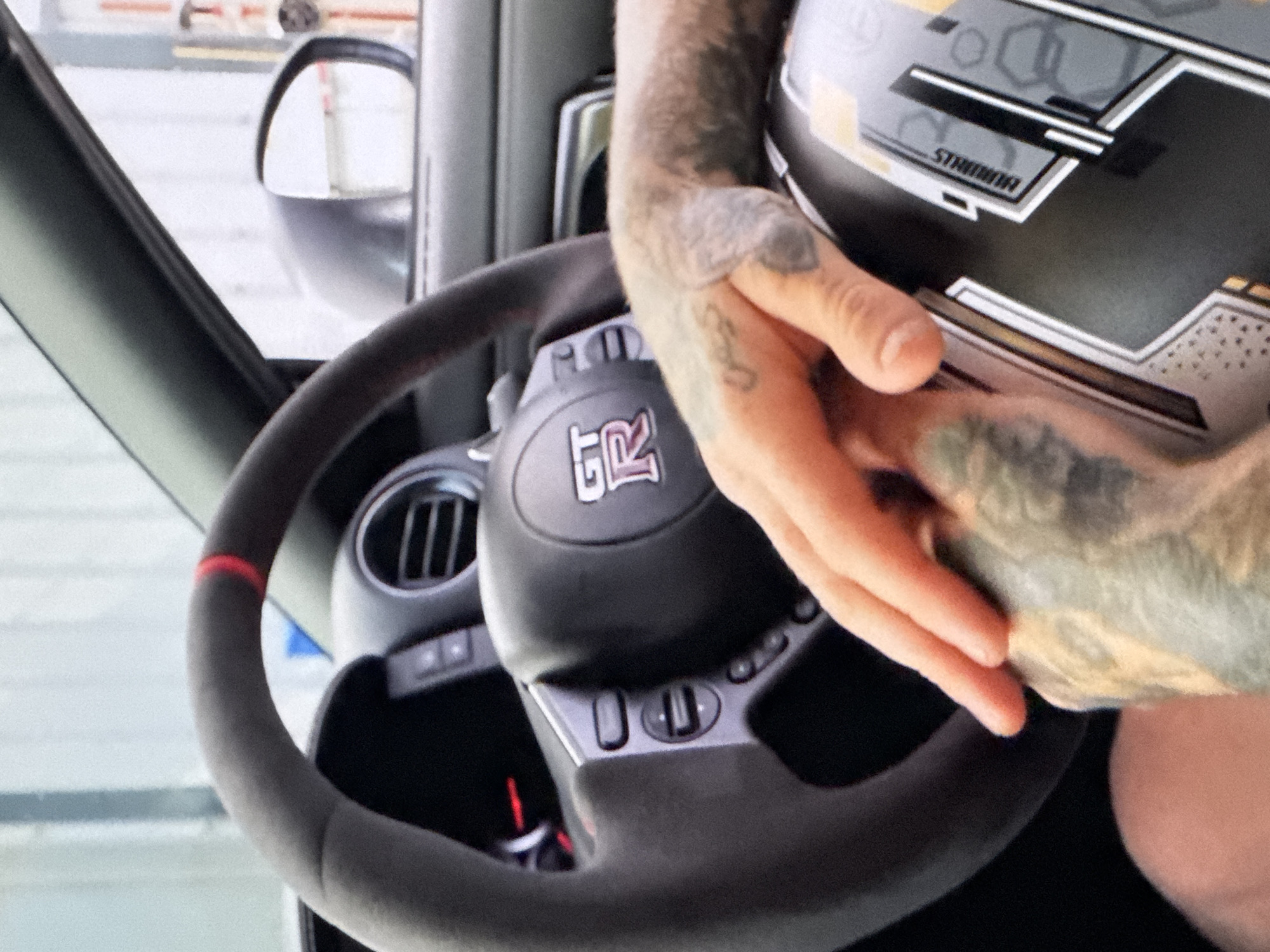 A person's hand, adorned with tattoos, rests on a dark helmet inside a vehicle. The helmet features a graphic design with the word "MINIMUS" visible. The steering wheel of the car, which has a red marking at the top, is also visible. The car's interior appears to be modern, with a visible air vent and dashboard controls. The background is blurred, suggesting the vehicle is either parked or in motion. The overall scene appears to be an indoor or outdoor setting, possibly a garage or a parking lot.